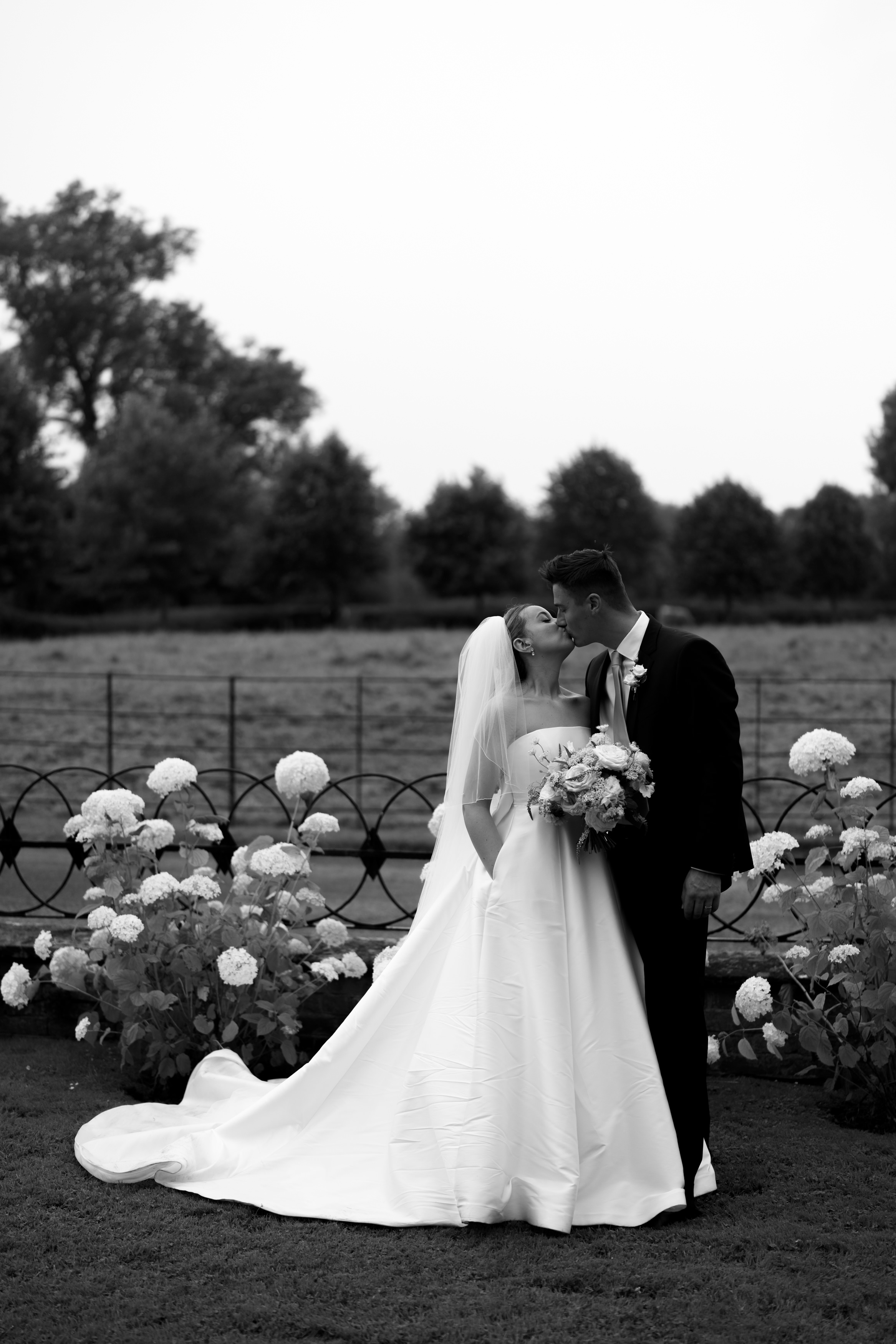 A bride and groom share a kiss outside, surrounded by hydrangea flowers. A wrought iron fence and trees can be seen in the background.