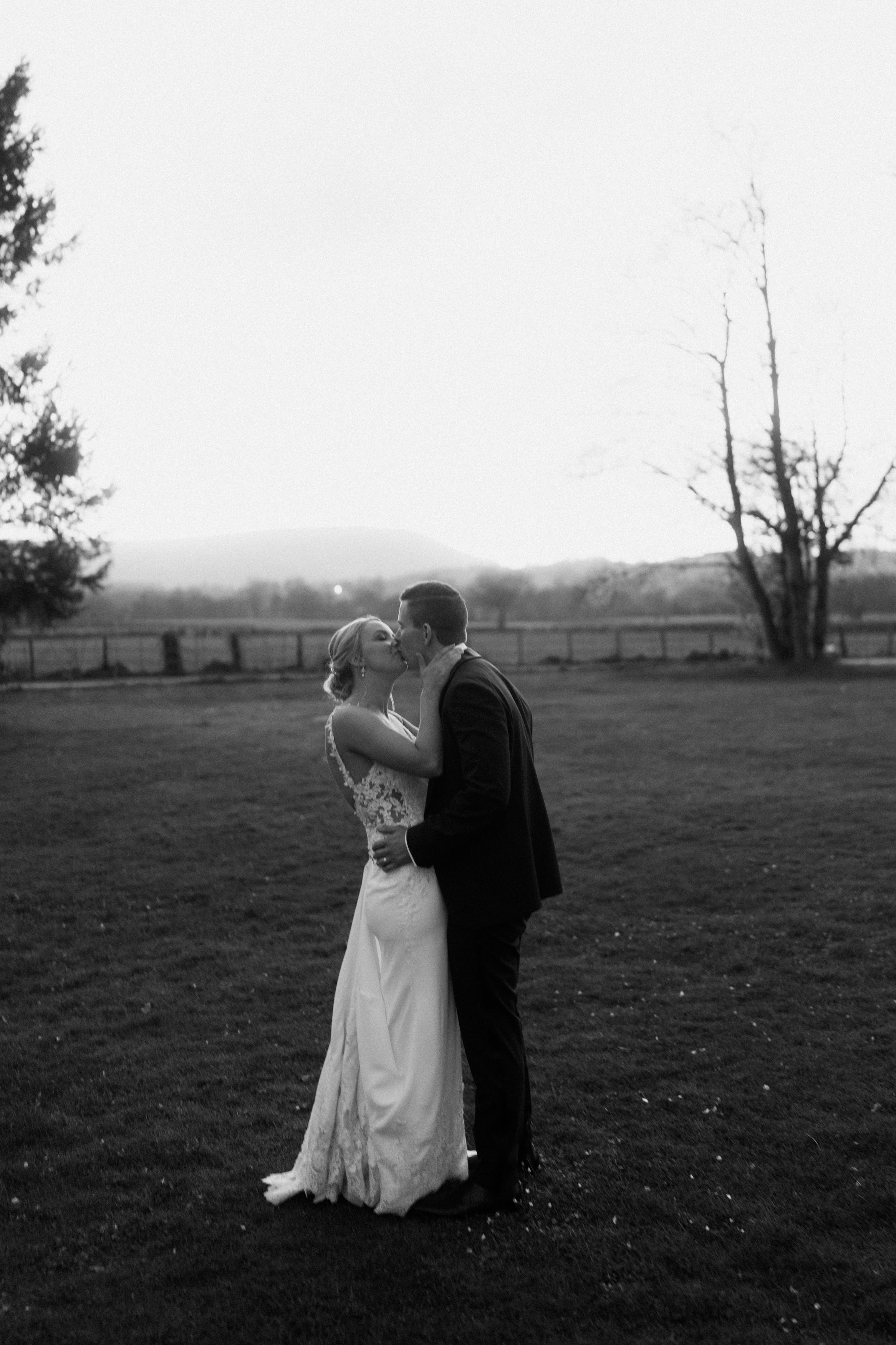A bride and groom kiss on a grassy field, with some trees and a fence in the background.