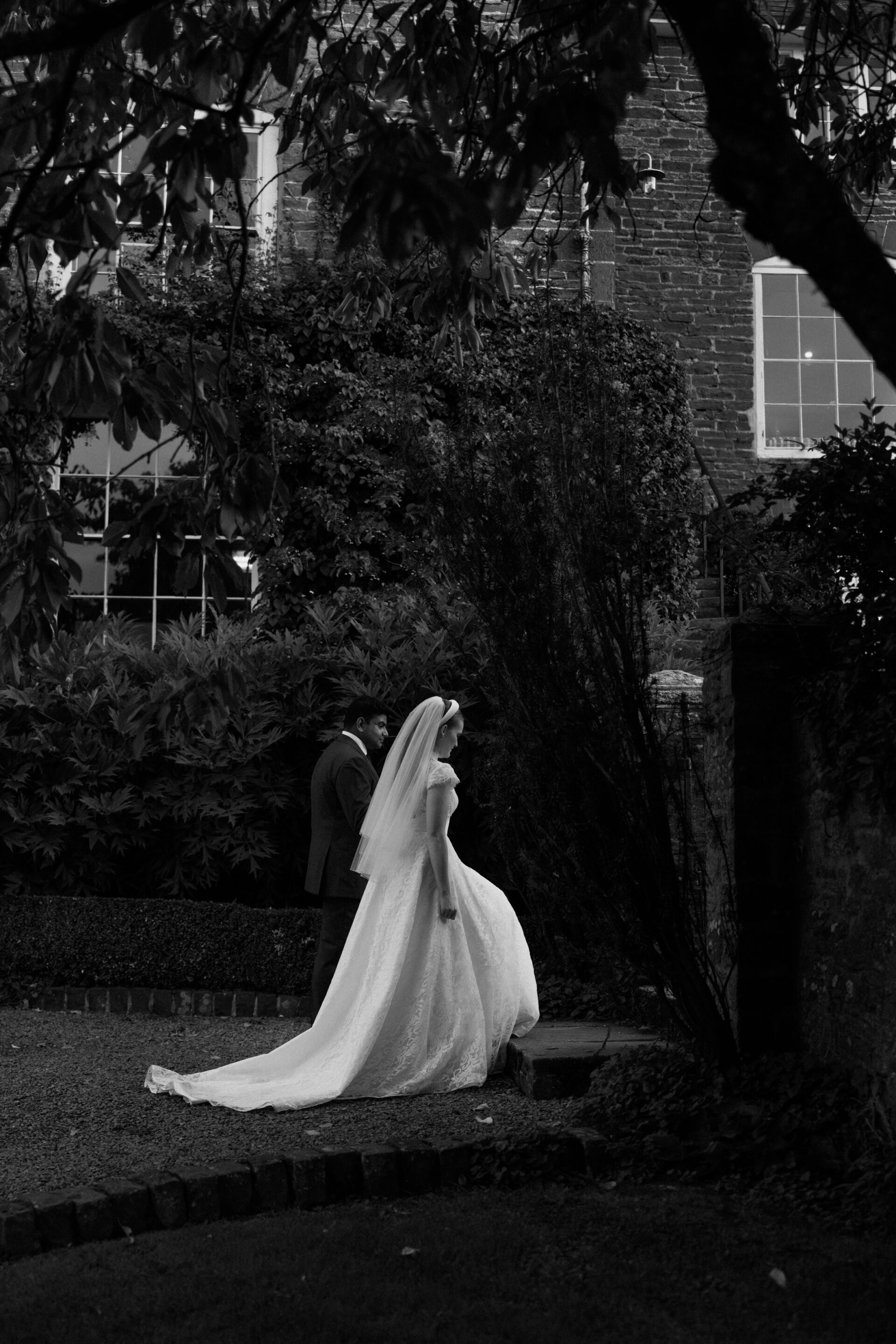 A bride wearing a wedding dress and veil walks outside next to a man in a suit, with trees and bushes by a brick building.