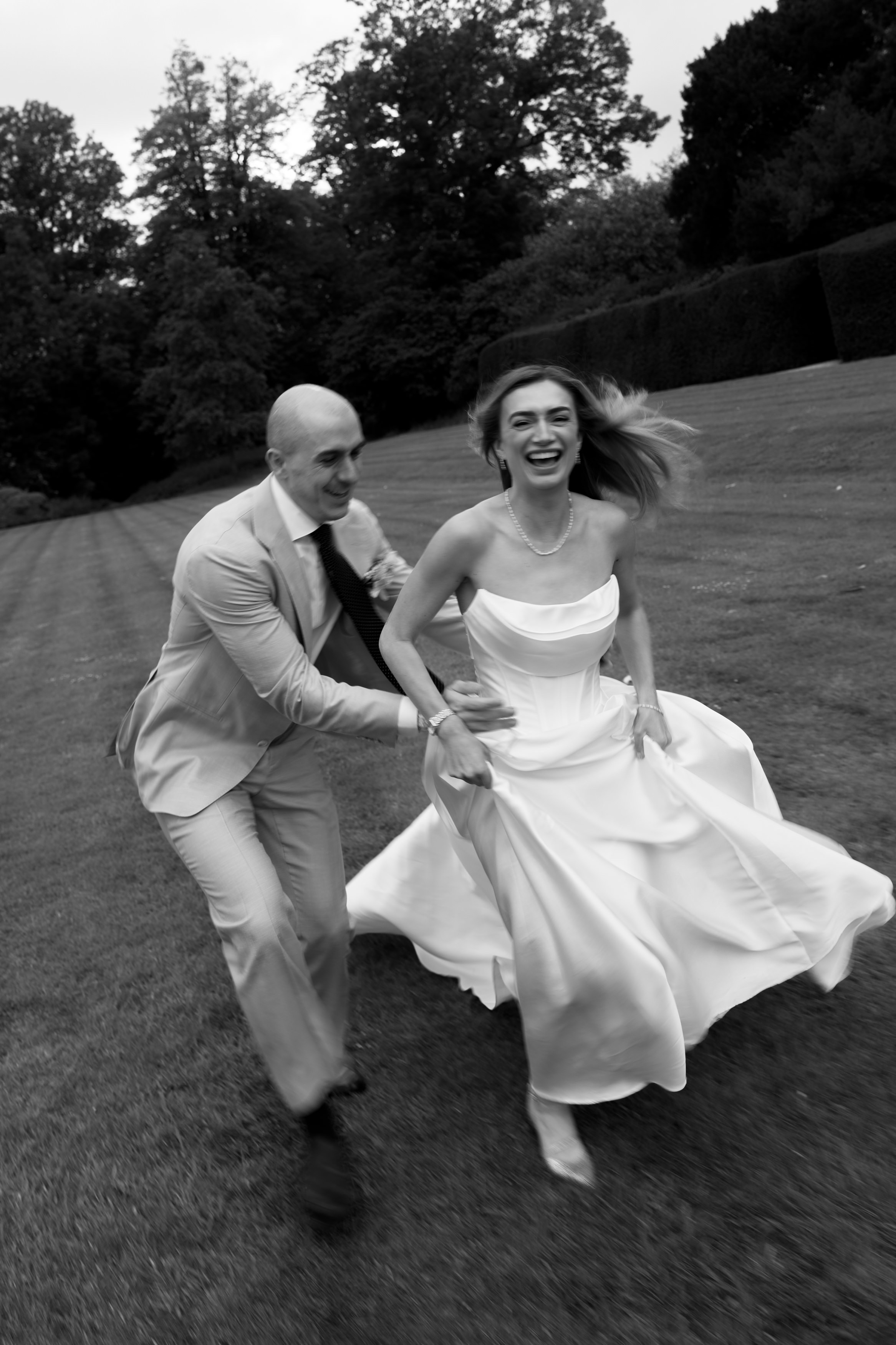 A man in a suit runs after a smiling woman in a white dress on the grass, with trees behind them. They both look happy.
