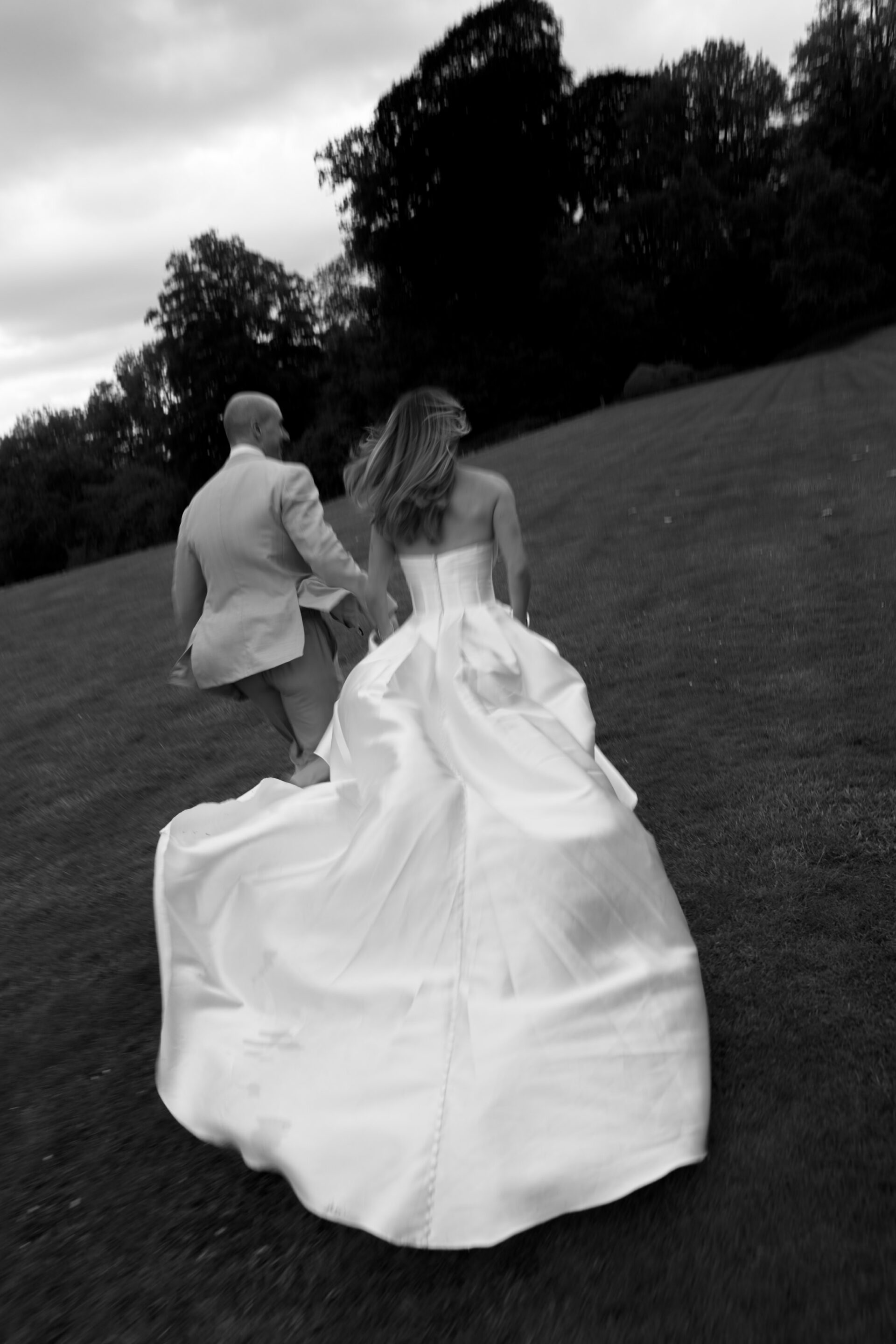 A woman in a wedding dress and a man in a suit walk together holding hands across a grassy field, shown in black and white.