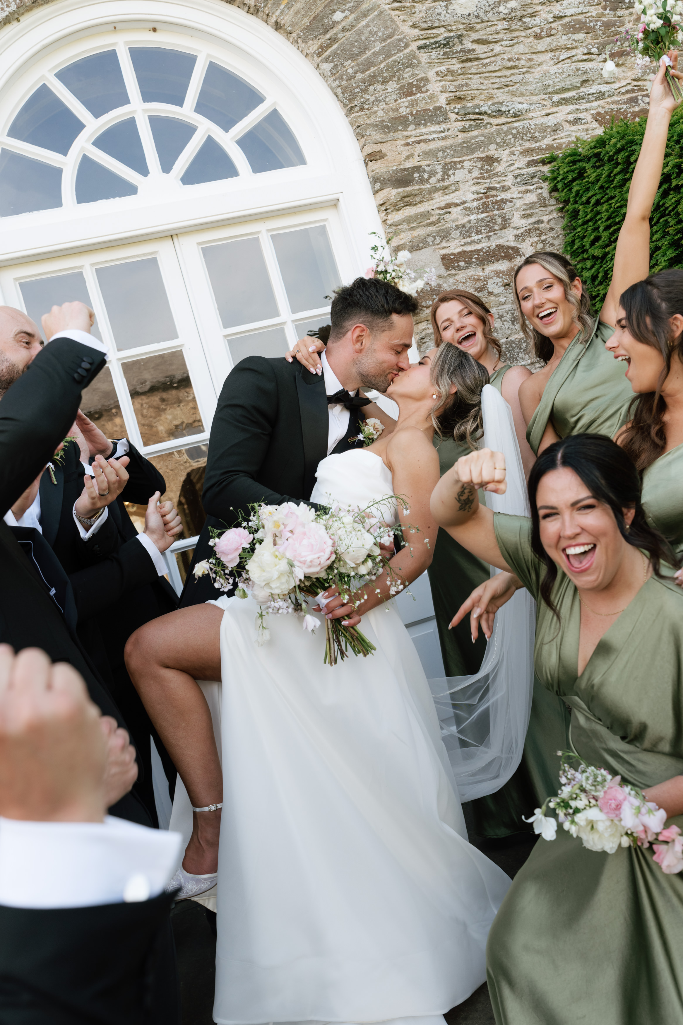 The bride and groom share a kiss outside while their happy, well-dressed friends cheer around them.