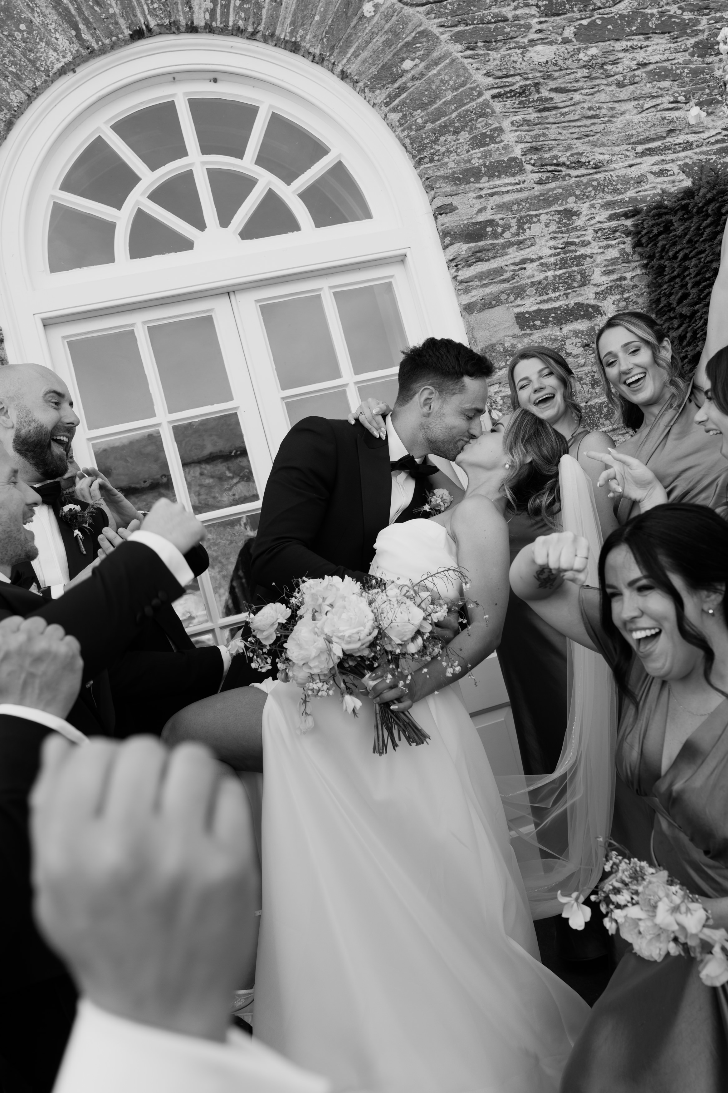 The bride and groom share a kiss holding a bouquet, whilst happy guests cheer outside a building with a big arched window.