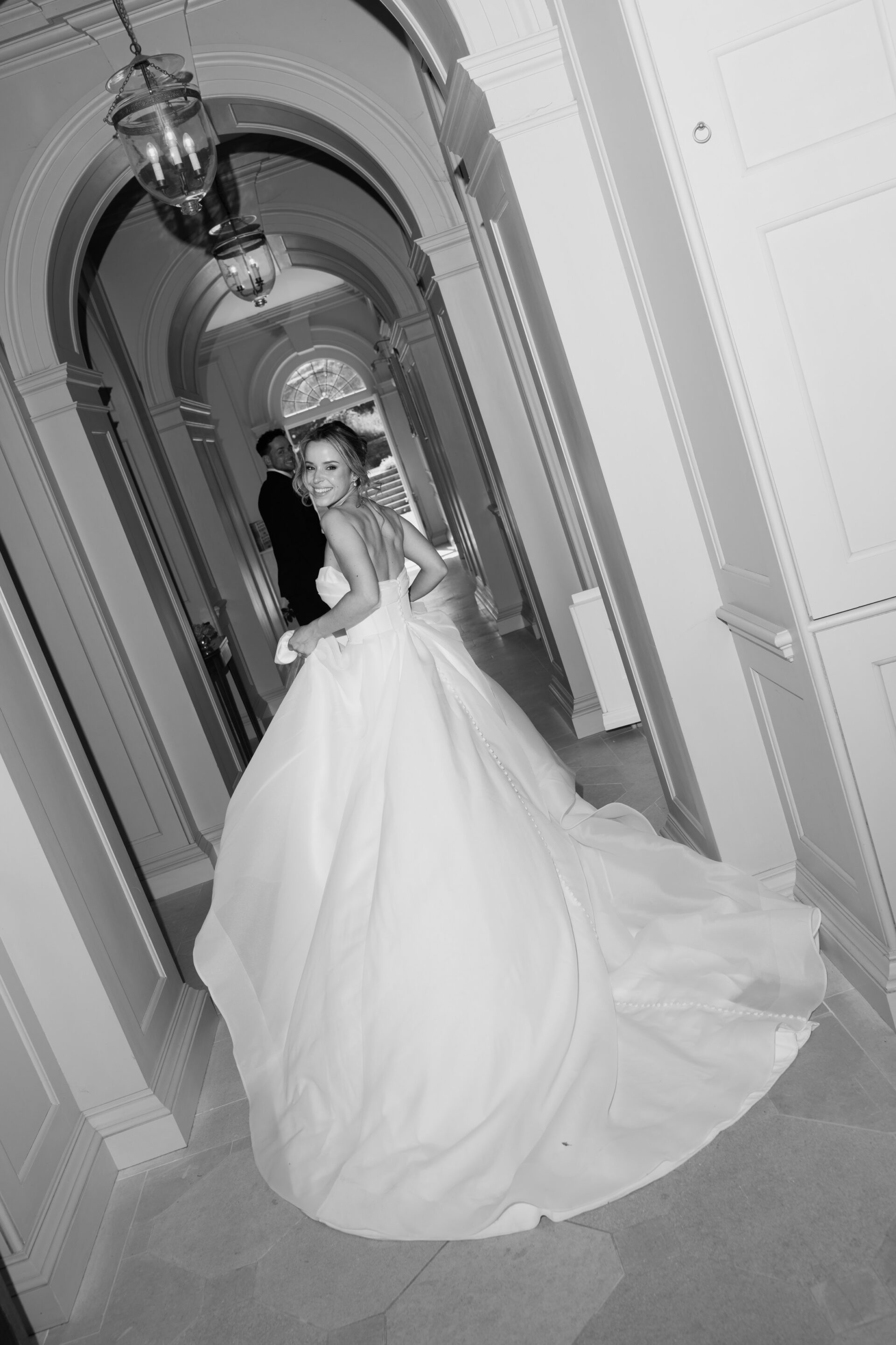 A bride in a strapless dress walks down a corridor, glancing back. A man in a suit is walking ahead. The photo is black and white.