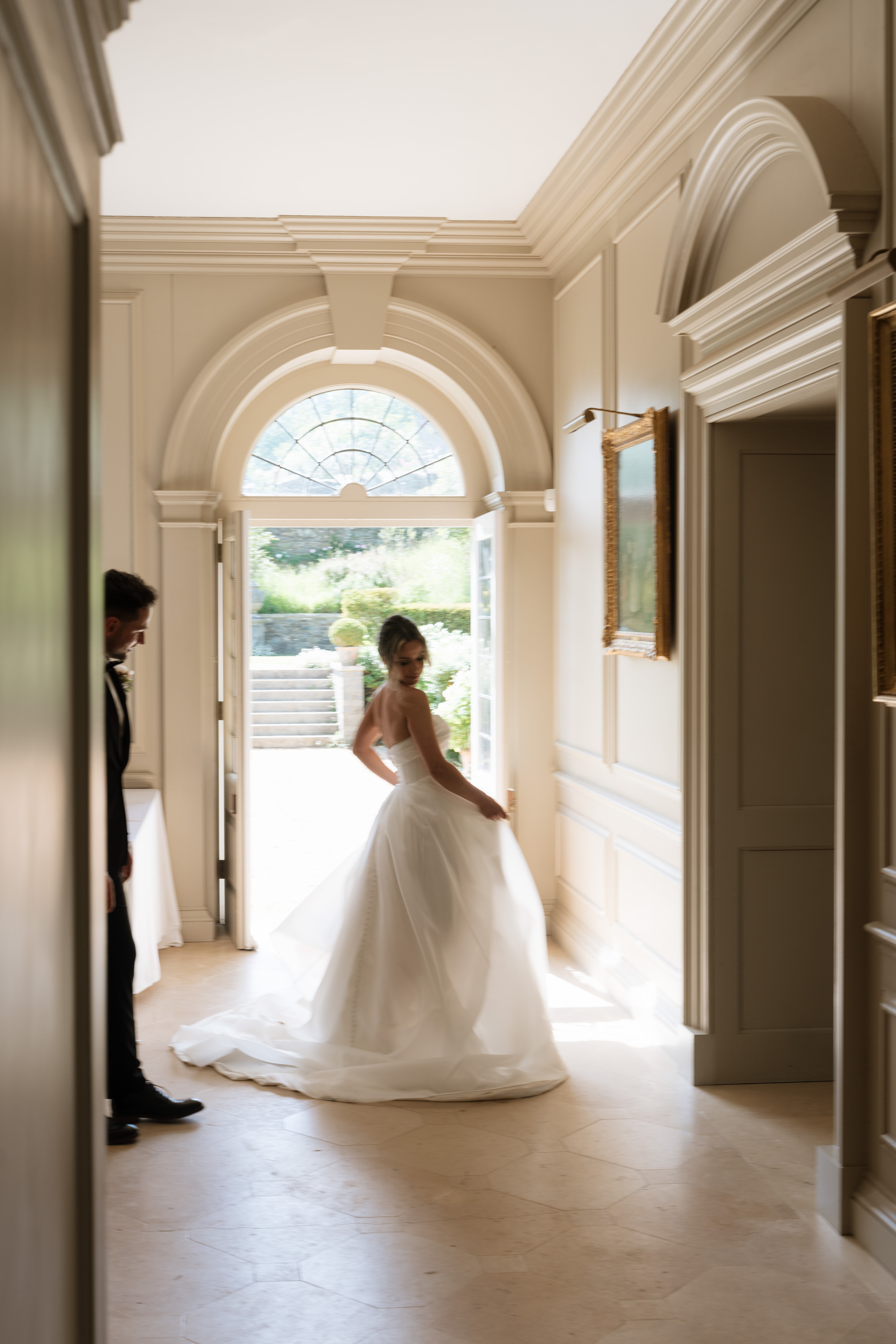 A bride in a white dress stands by a sunny doorway, glancing back, with a man in a suit partly hidden in the shadows close by.