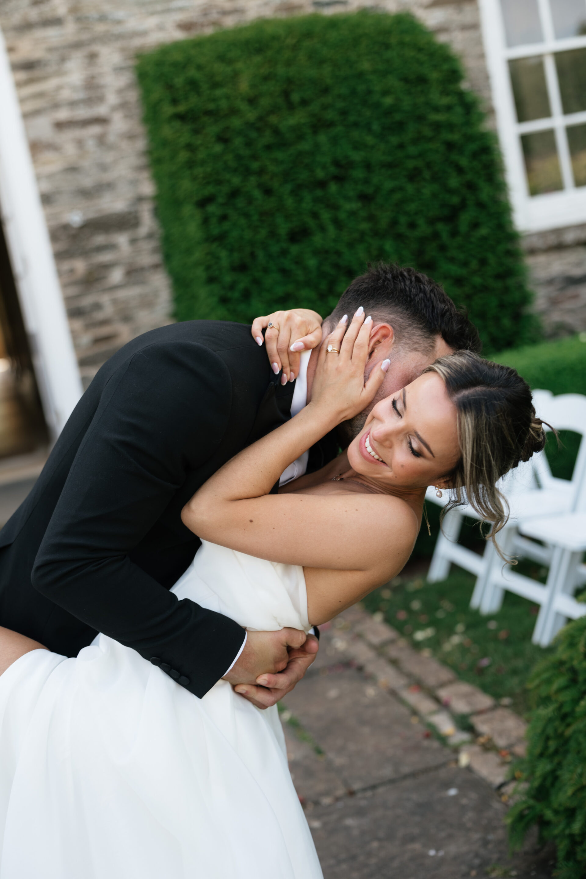 A bride and groom hug and smile outside. The groom bends down as the bride holds his head. White chairs and greenery are behind them.