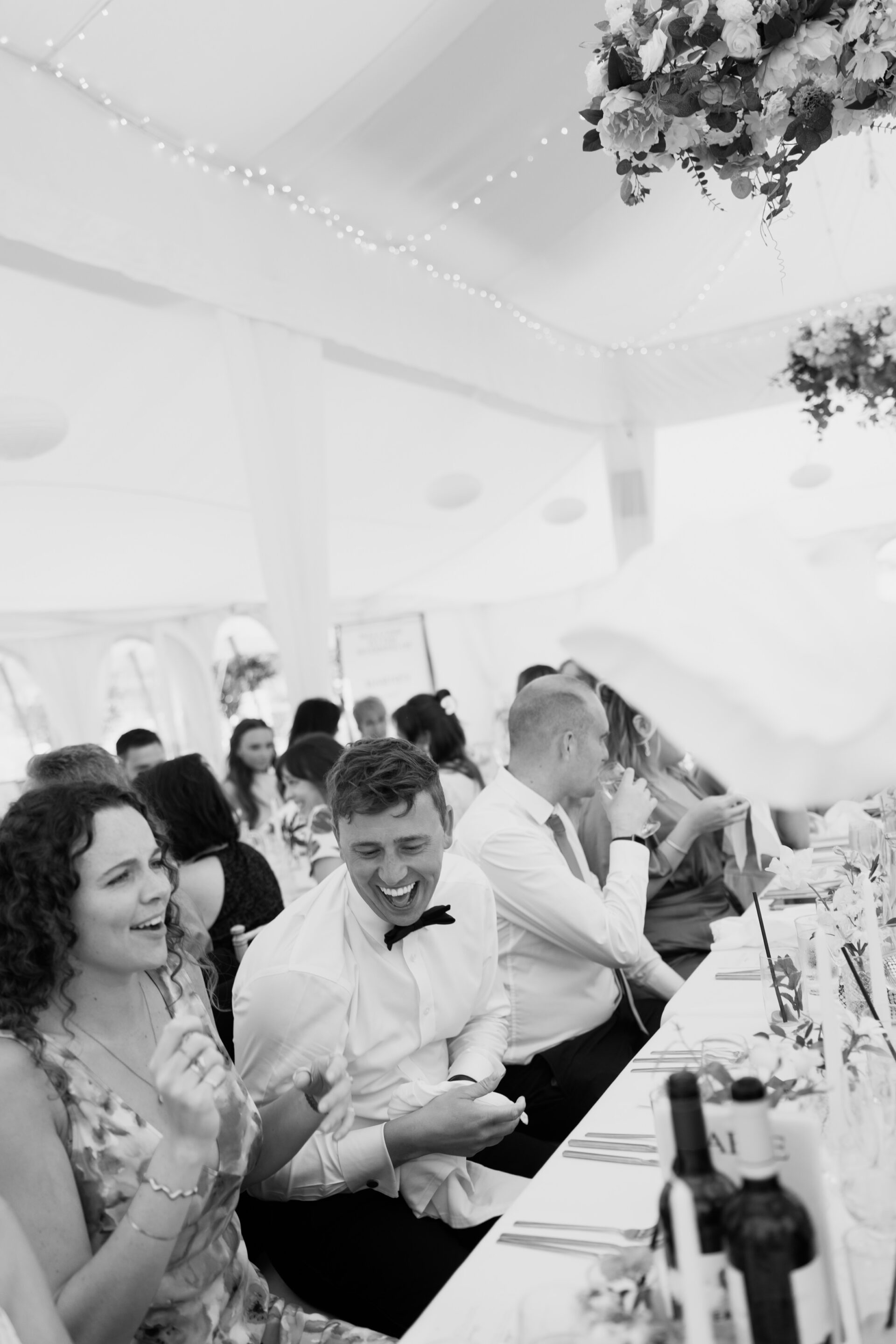 People sitting at a long table in a decorated marquee, enjoying a formal event with flowers and wine bottles on the table.