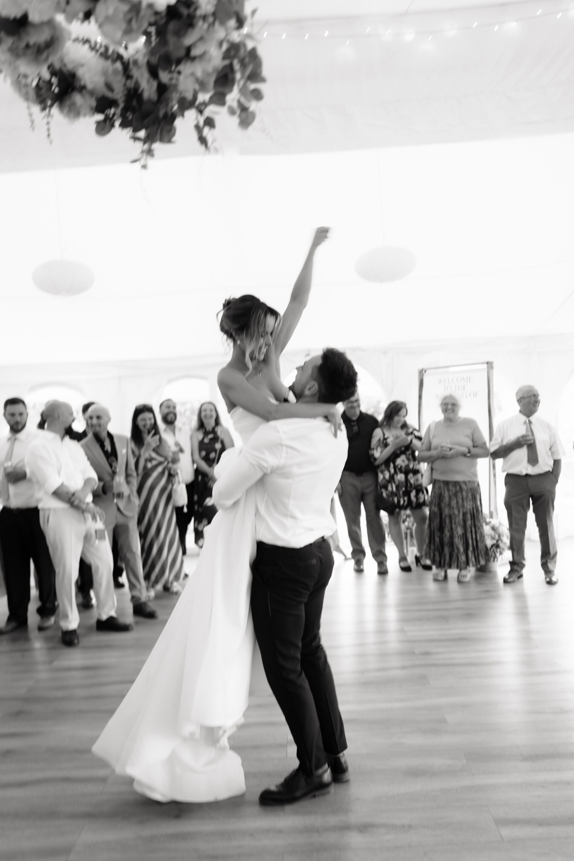 The groom lifts the bride whilst they dance, and their guests watch and smile in the background.