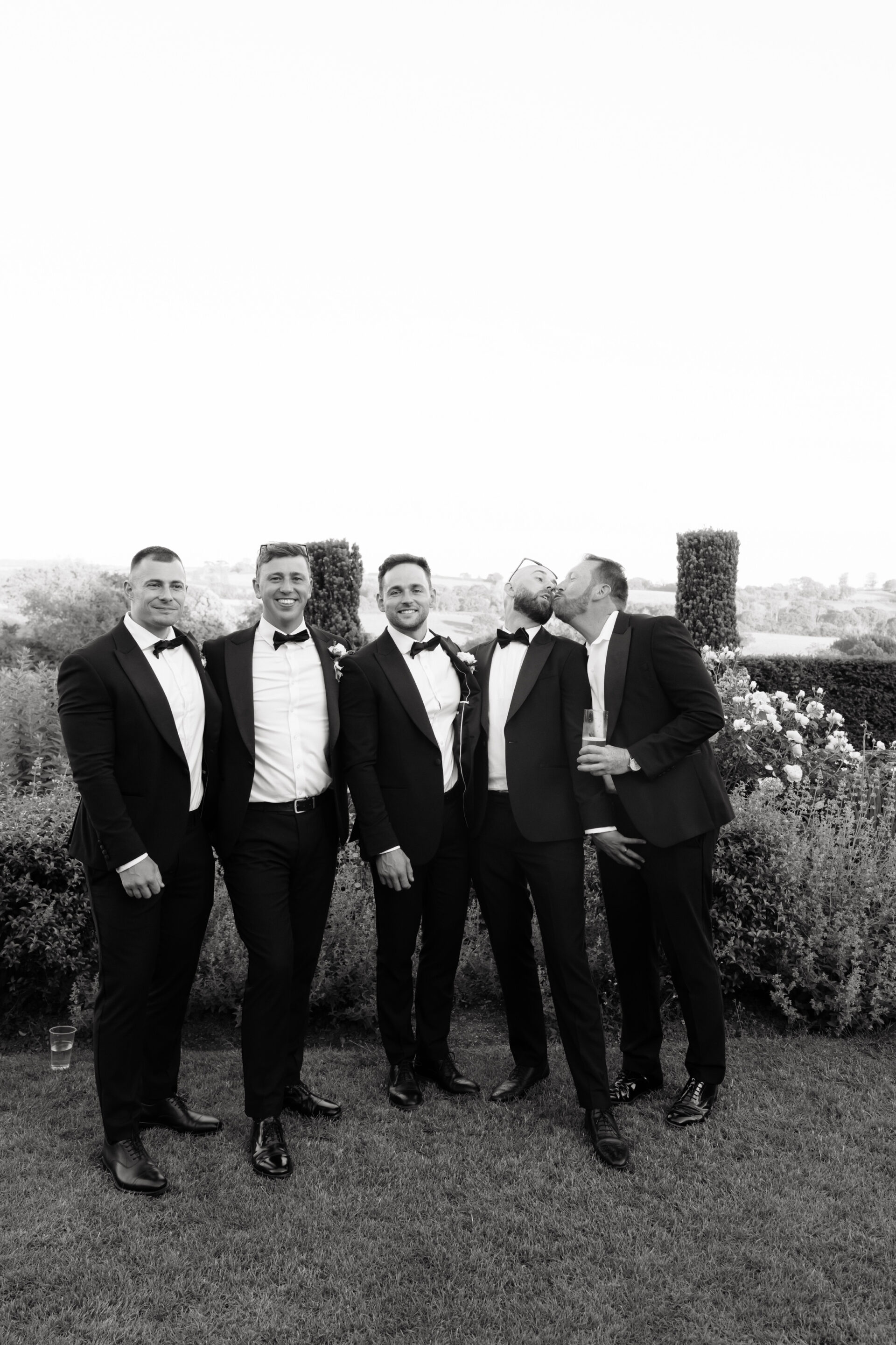 Five blokes in suits stand outside; one kisses another on the cheek while the rest smile and pose for a photo.