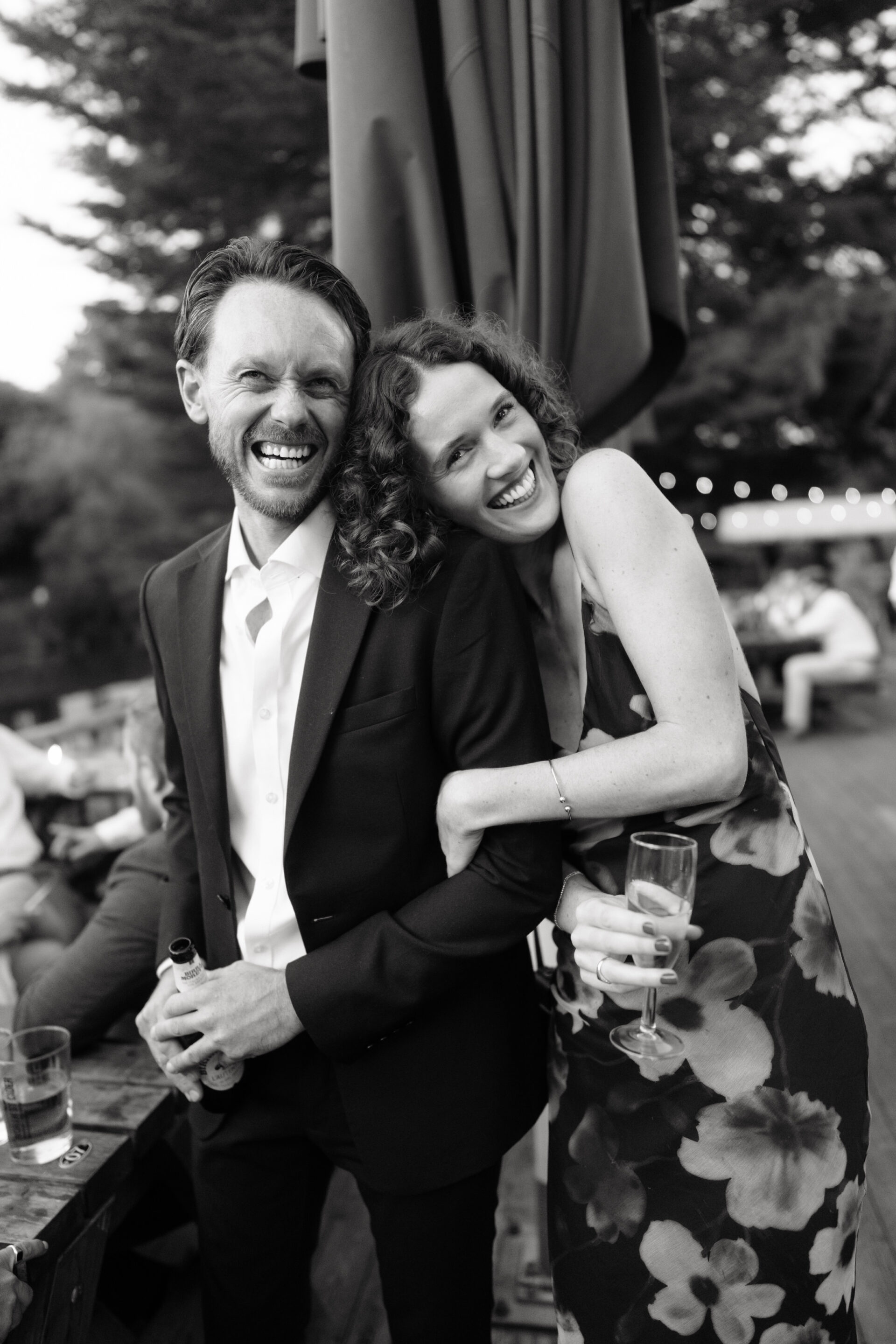 A man wearing a suit and a woman in a floral dress smile as they stand close together outside, both holding drinks.