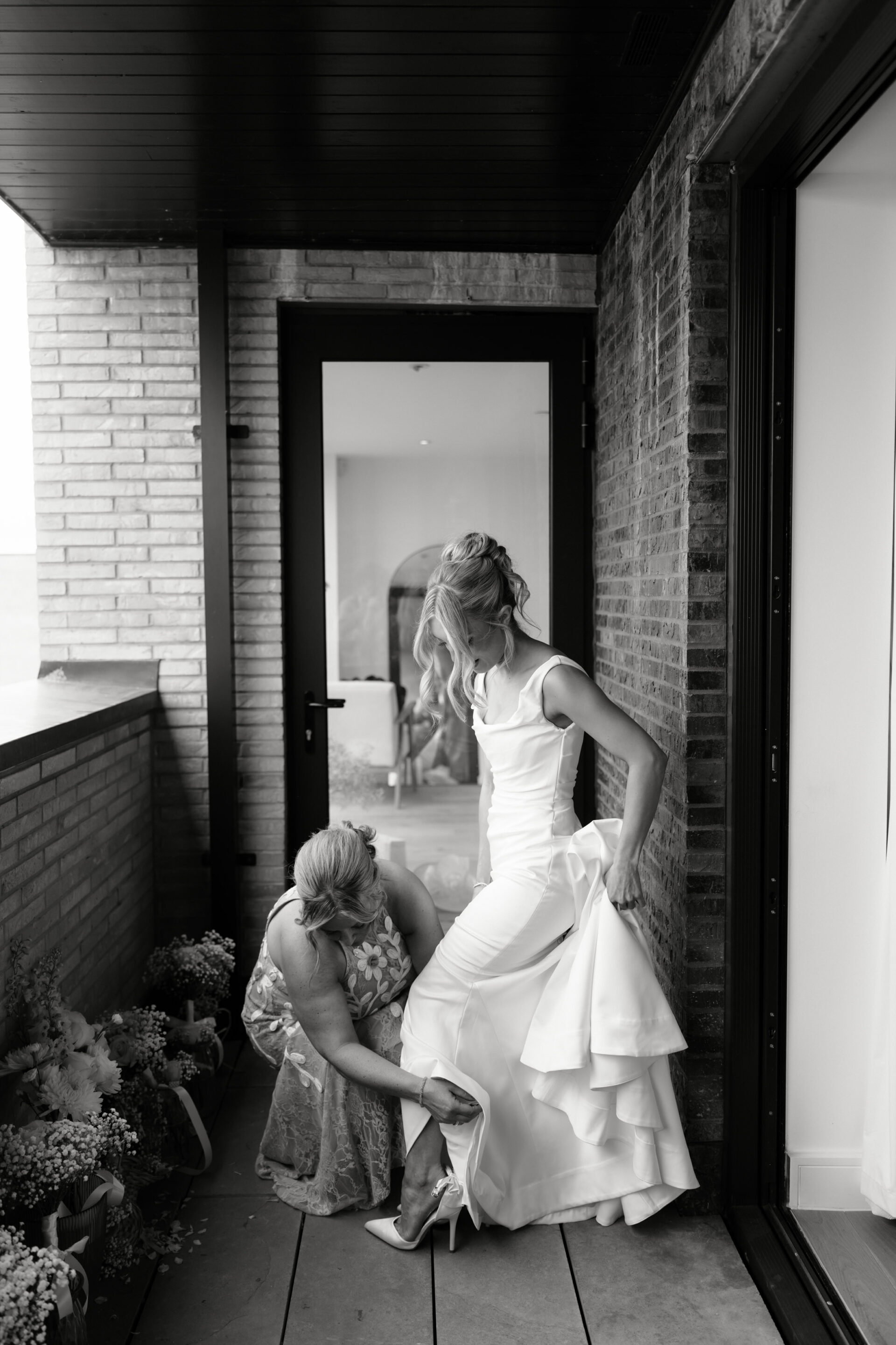 A bride stands on a balcony as another woman helps her put on her shoe. There are flowers lined up along the floor.