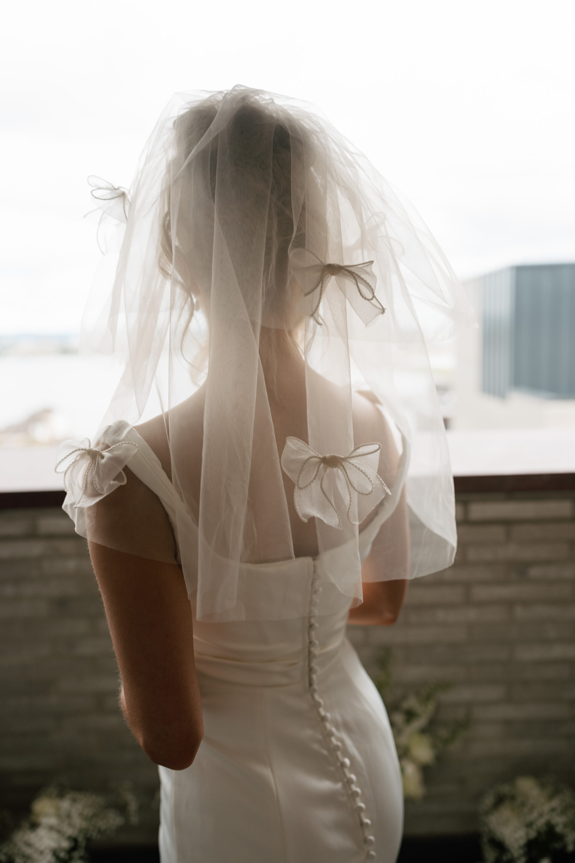 A bride in a white dress with a veil stands on a balcony, her back to the camera, and buttons lining the back of her gown.