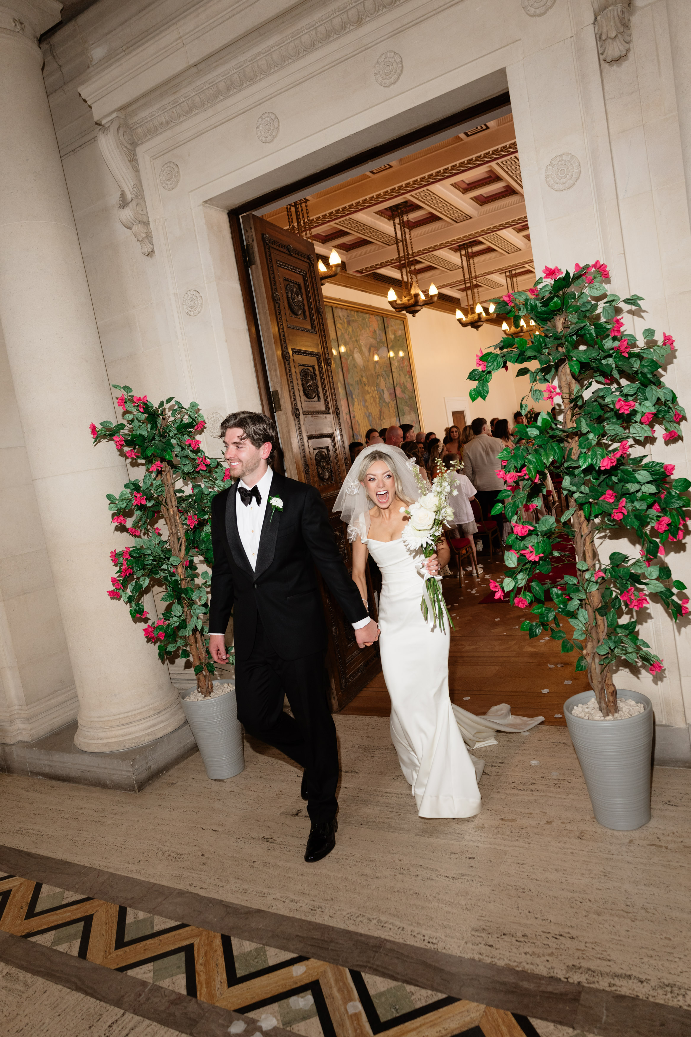 A bride and groom, dressed up, walk out of a decorated door holding hands and smiling, with guests behind them.