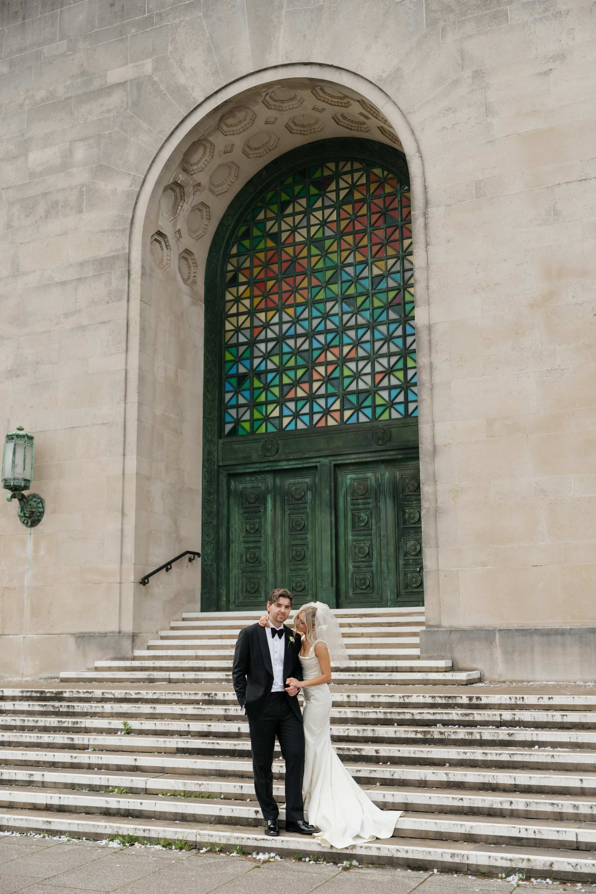 A bride and groom are standing side by side on stone steps in front of a big green door with brightly coloured stained glass overhead.