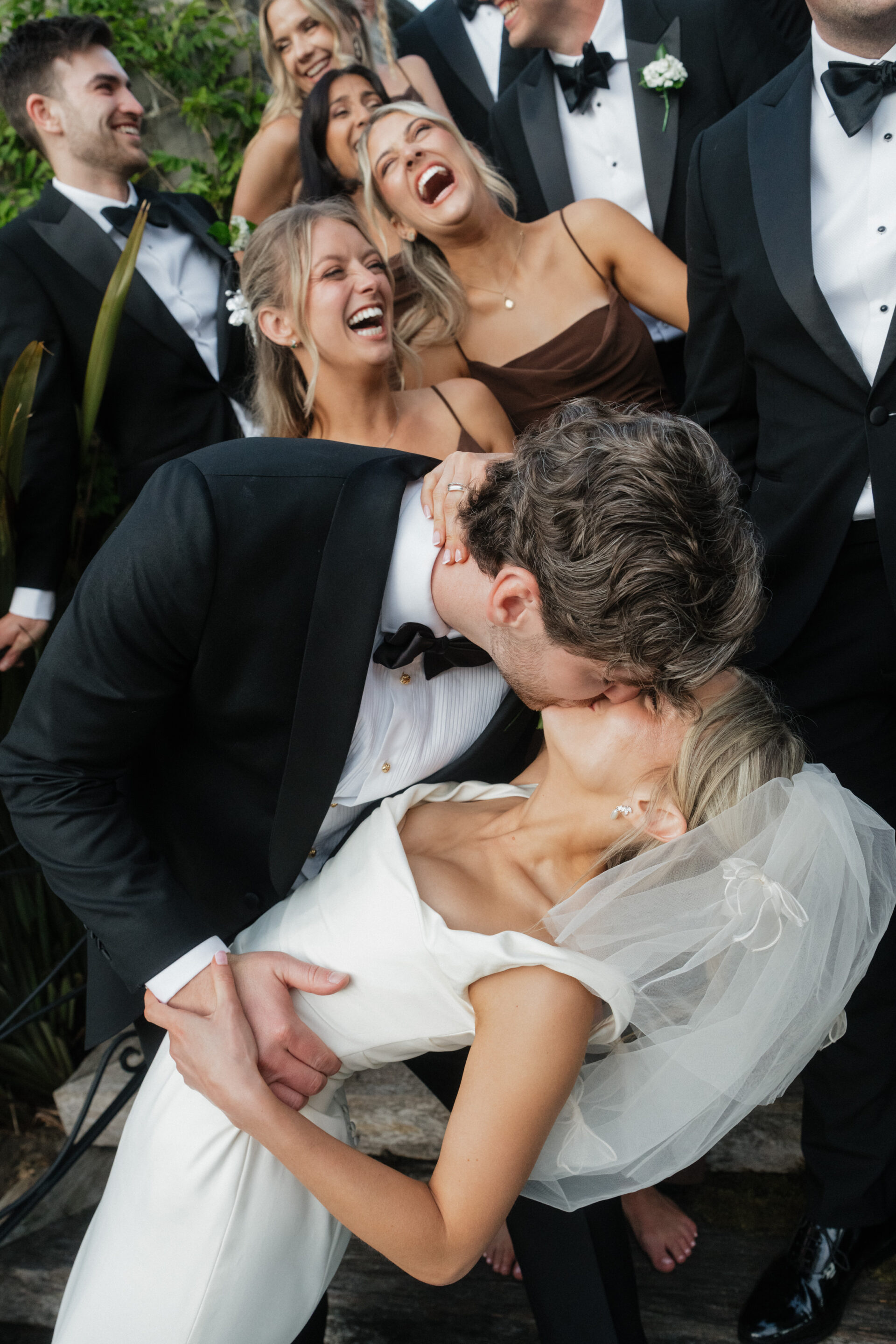 A groom in a black dinner suit dips and kisses his bride, who’s wearing a white dress and veil, whilst their wedding party smiles behind them.