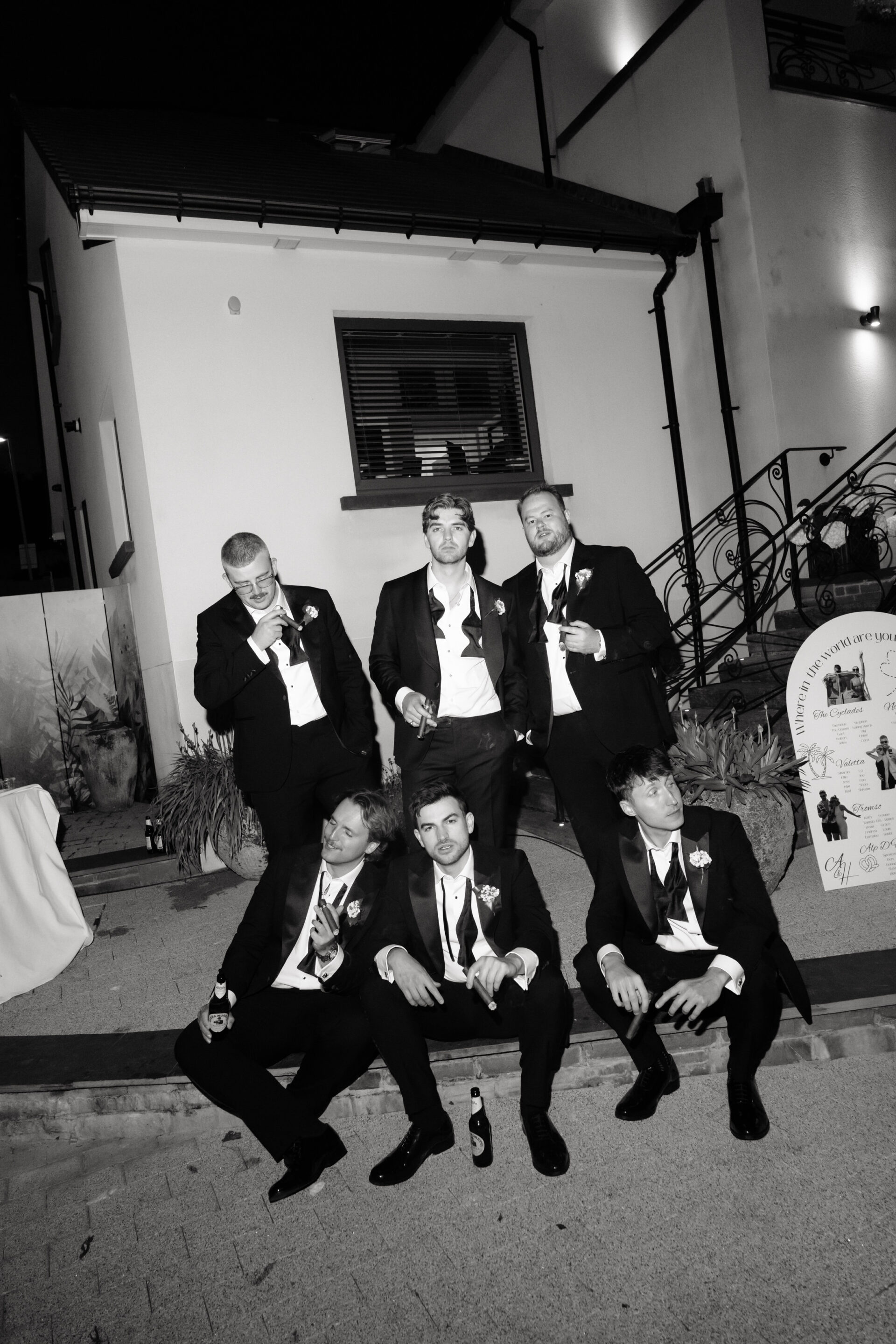 Six men in suits gather outside a building at night for a group photo, some standing and some sitting, holding drinks and cigarettes.