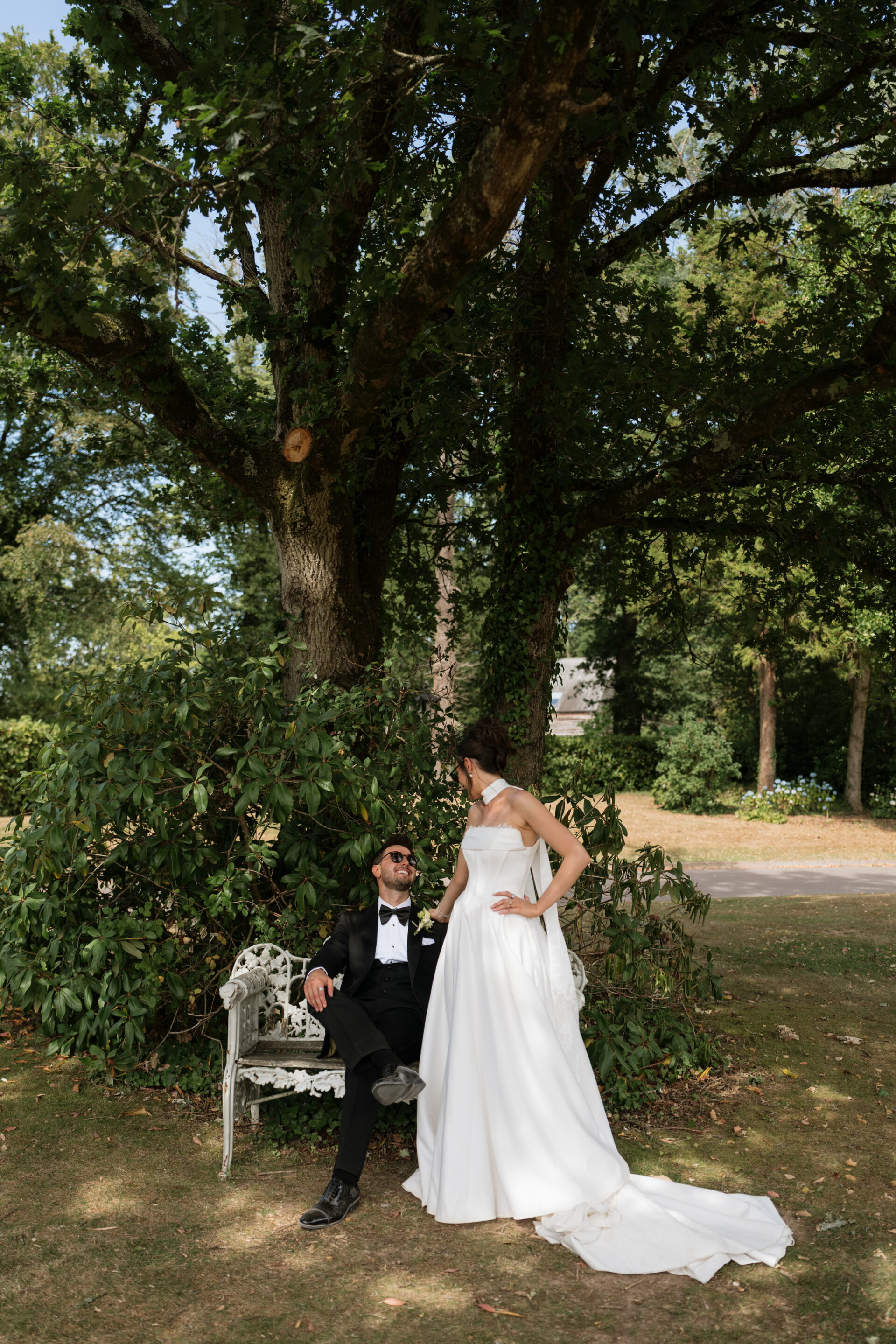 A bride wearing a white dress stands beside a groom in a dinner suit who’s sitting on a bench under a large tree in an outdoor garden.