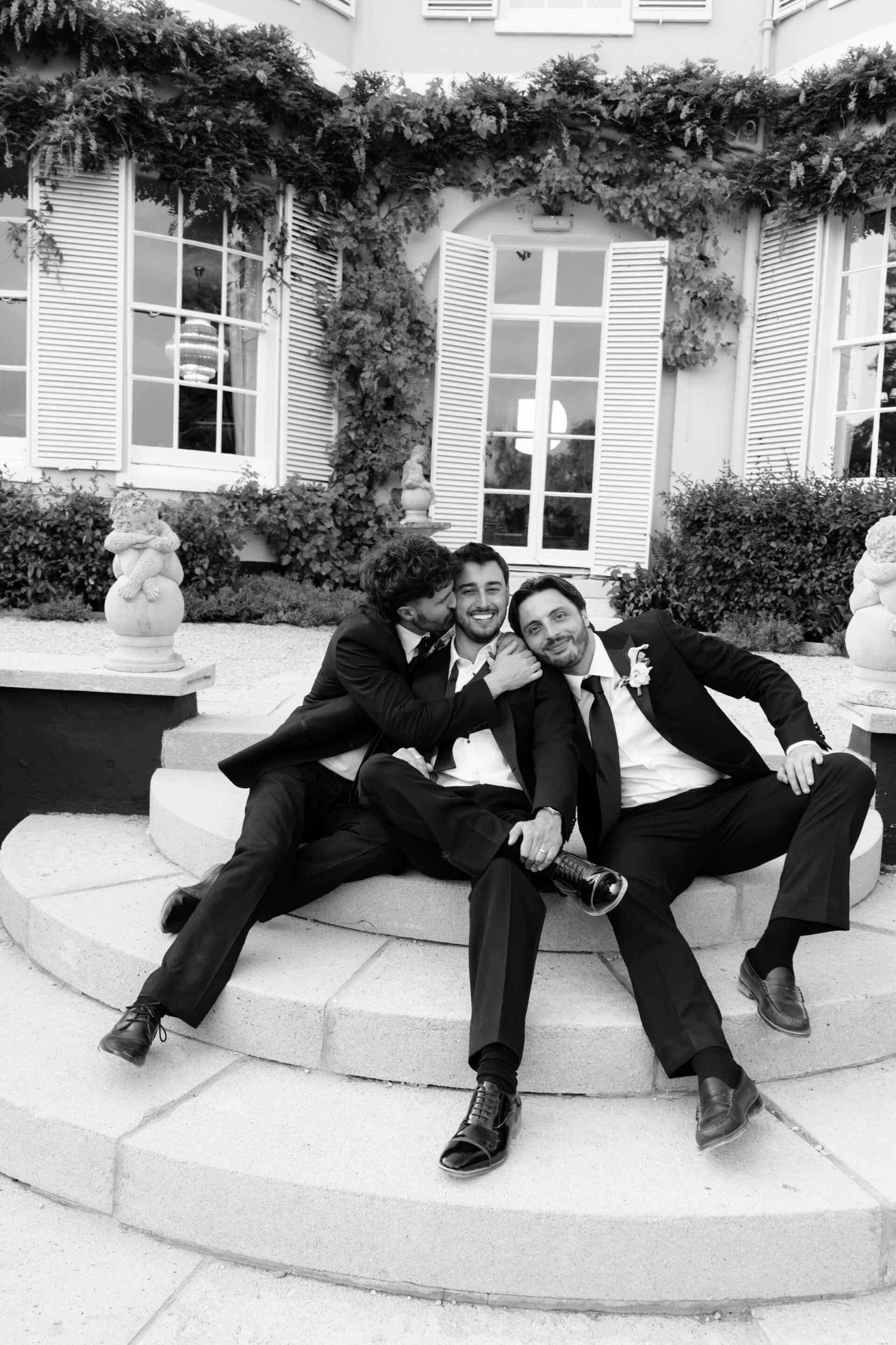 Three men wearing suits are sitting on steps outside a building, smiling for a photo. Two of them lean in towards the bloke in the middle. They all look happy and relaxed.