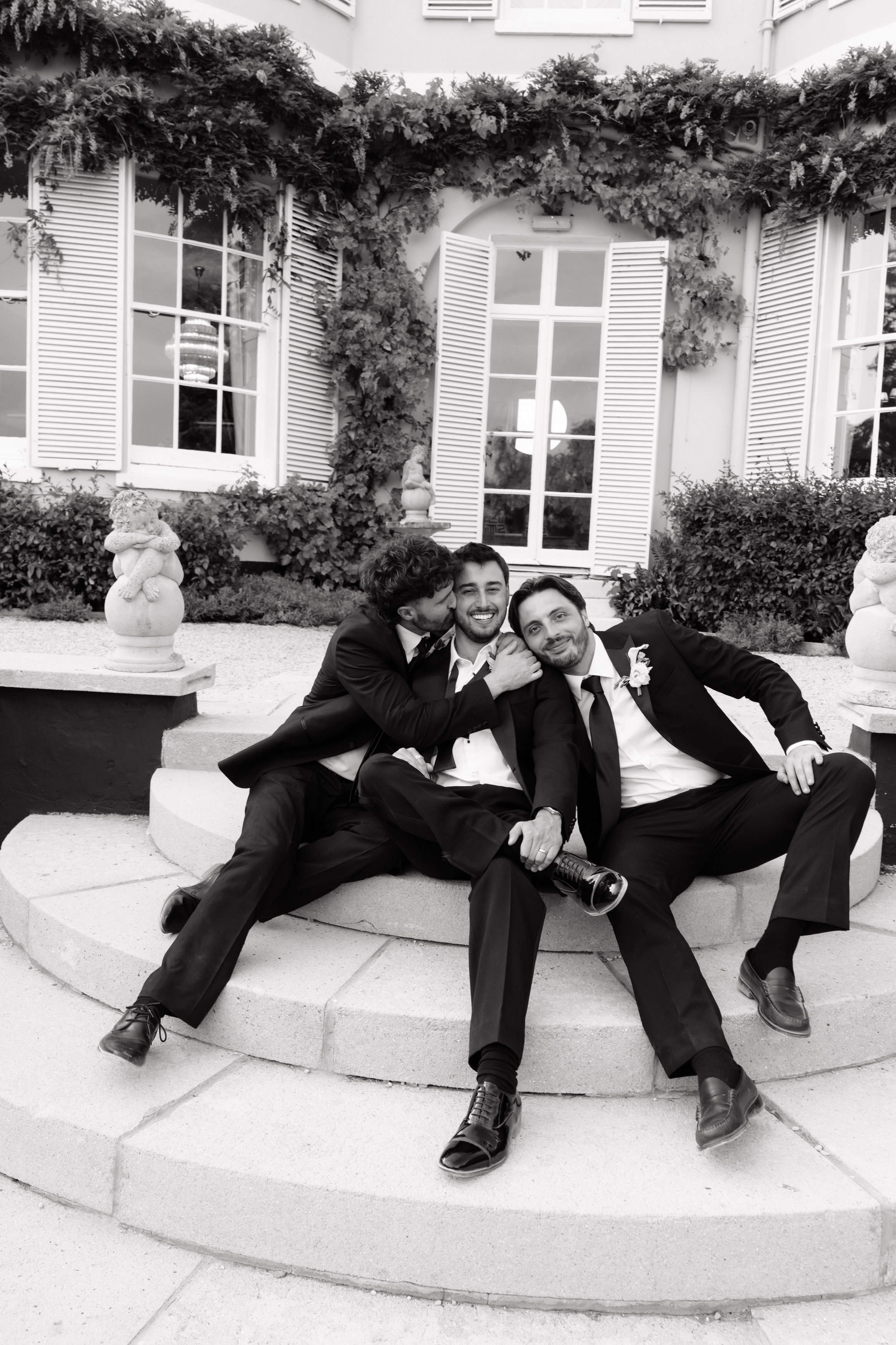 Three men wearing suits are sitting on steps outside a building, smiling for a photo. Two of them lean in towards the bloke in the middle. They all look happy and relaxed.