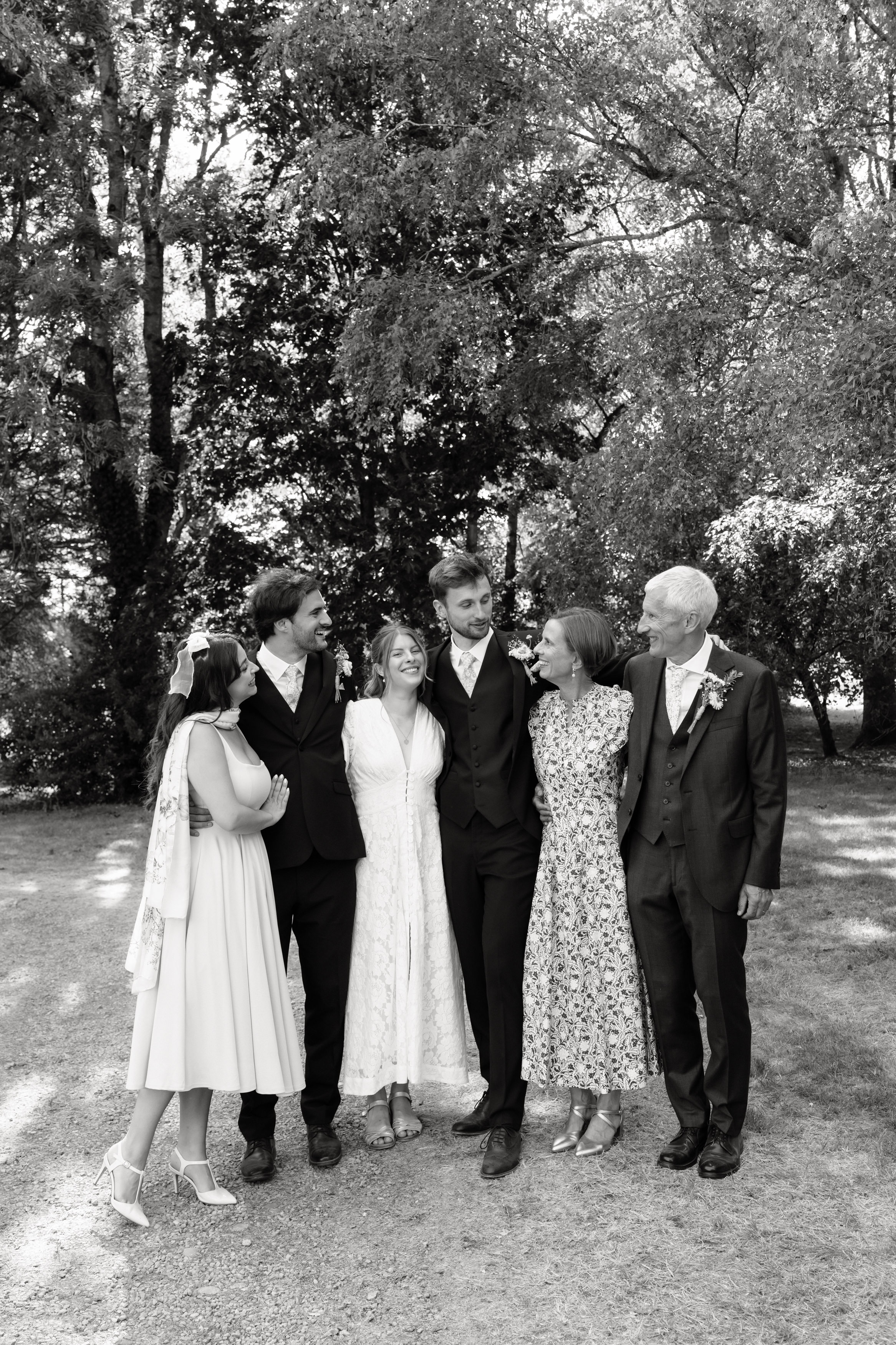 Six people, dressed up nicely, are standing together in a park. They’re smiling and looking at one another. There are three men and three women, with trees around them and sunlight shining through.