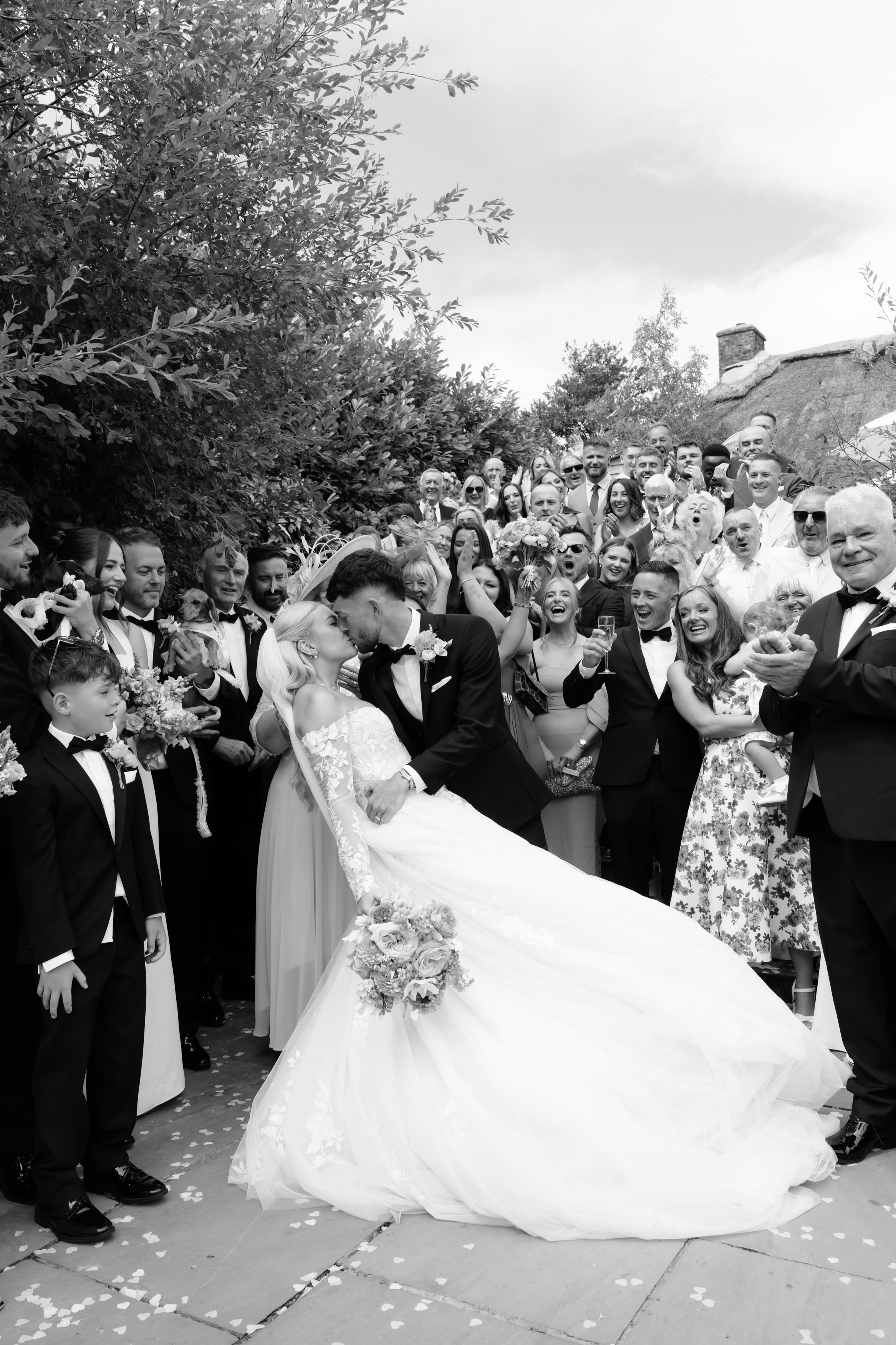 A bride and groom share a kiss outside as their guests smile and clap around them during the wedding.