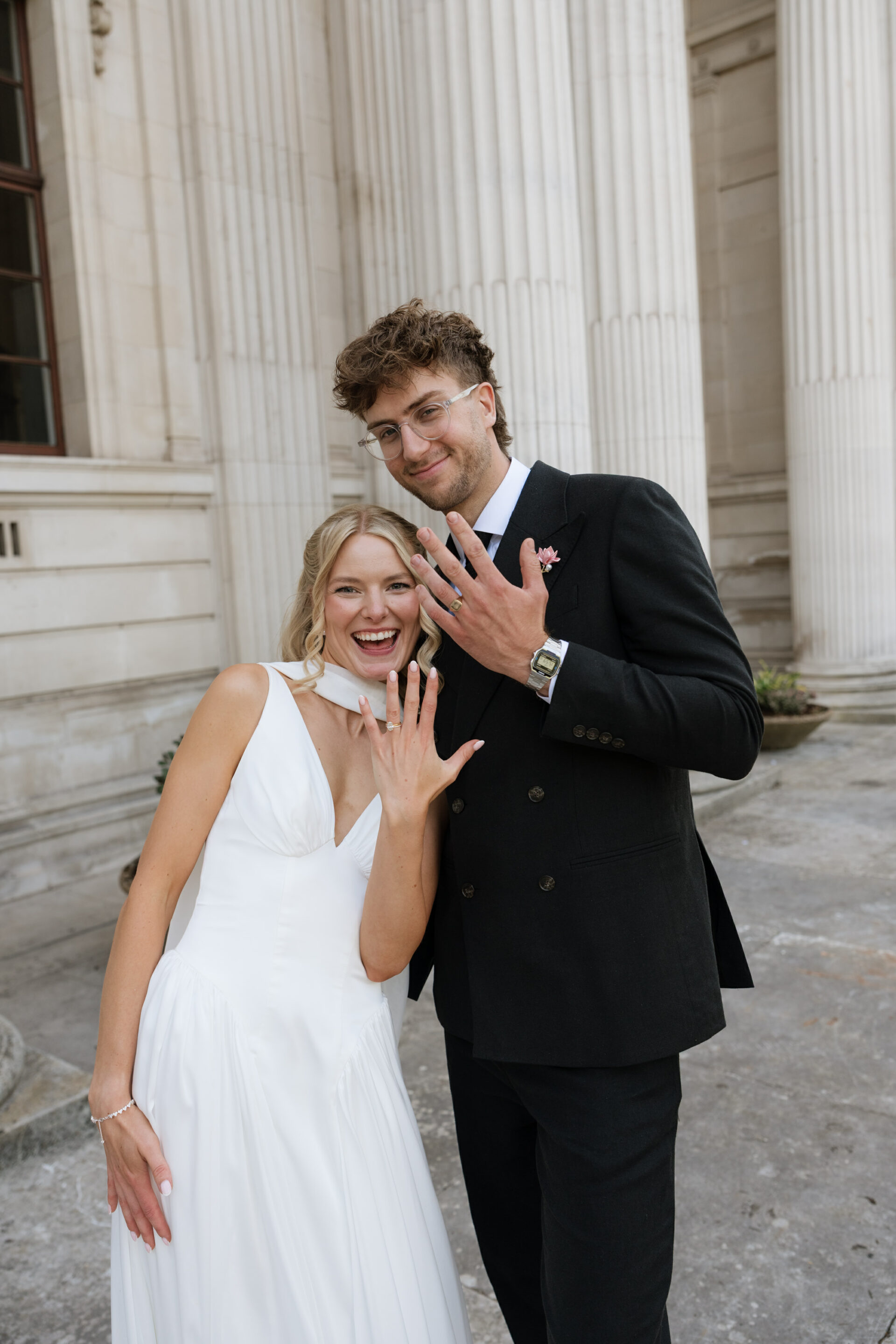 A bride wearing a white dress and a groom in a black suit are standing outside a building. They’re both smiling and holding up their hands to show their rings to the camera.