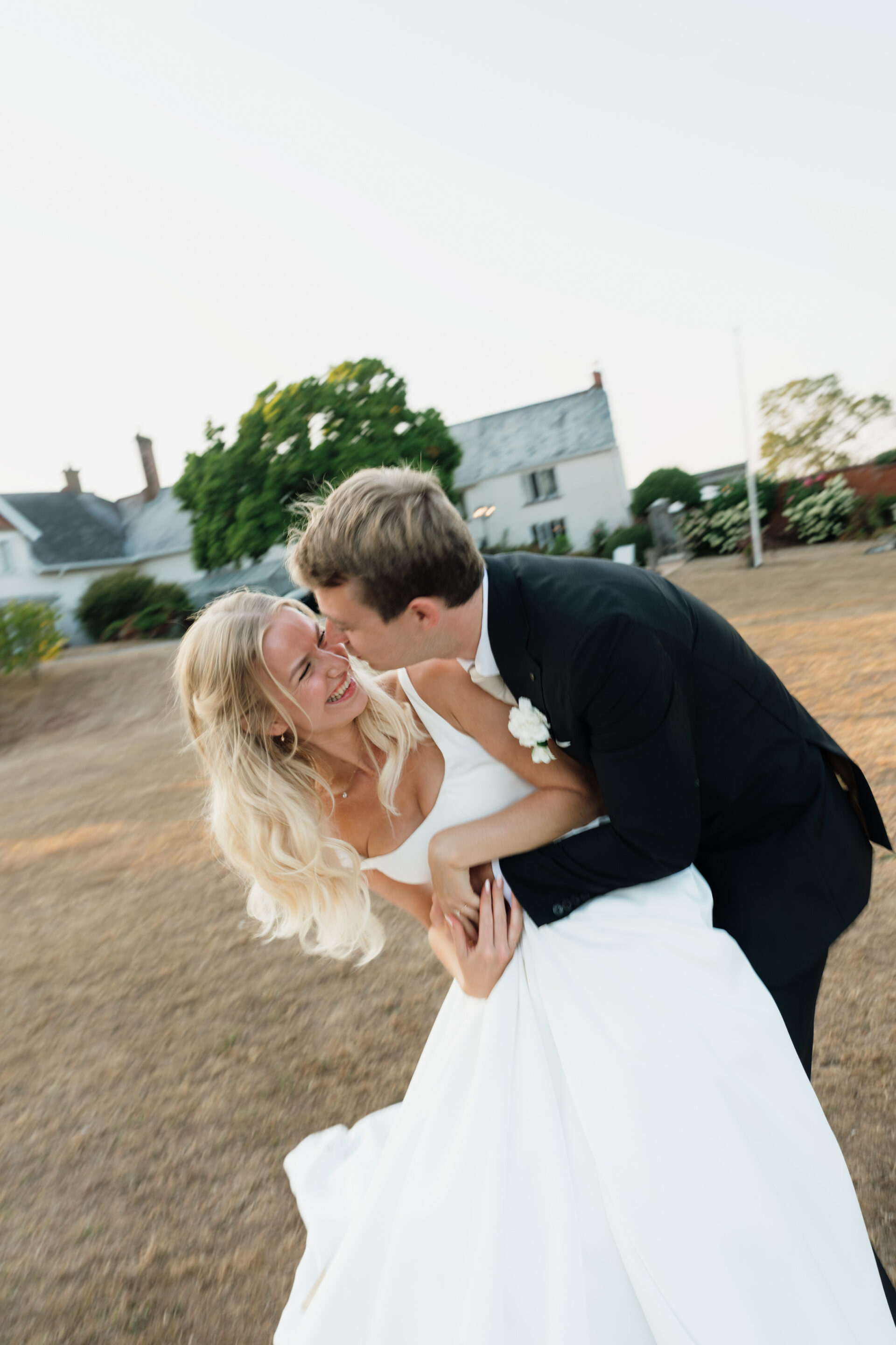 A bride and groom are laughing and hugging each other outside on a patch of dry grass, with some houses and trees in the background.