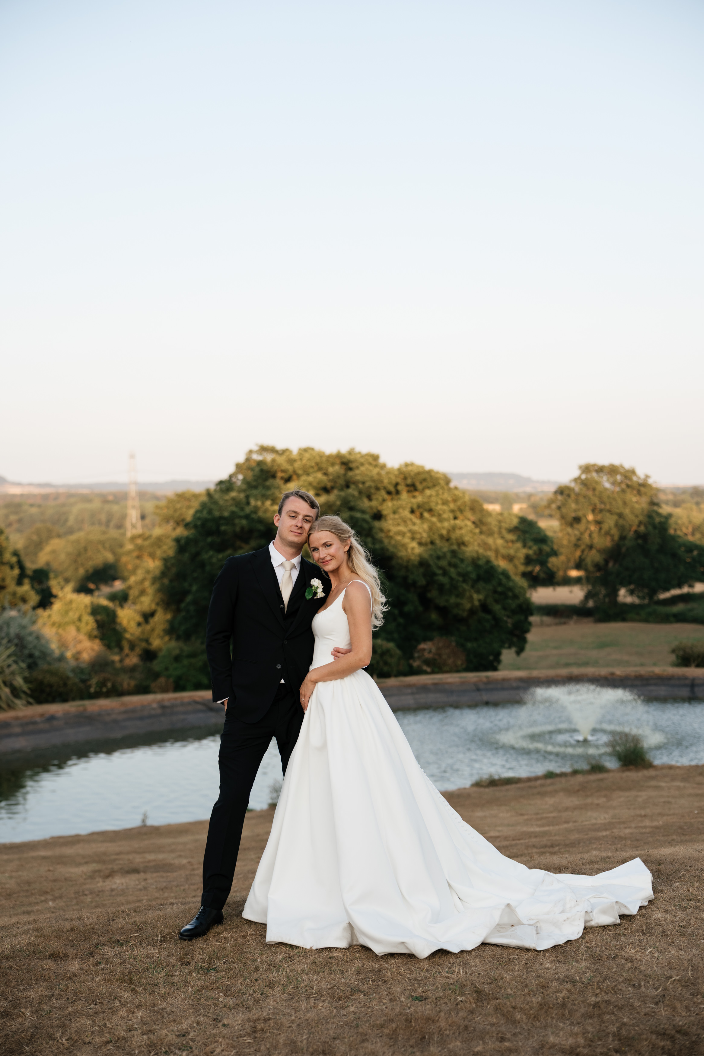 A bride wearing a white dress and a groom in a black suit are standing outside on some brown grass, with a pond and trees behind them.