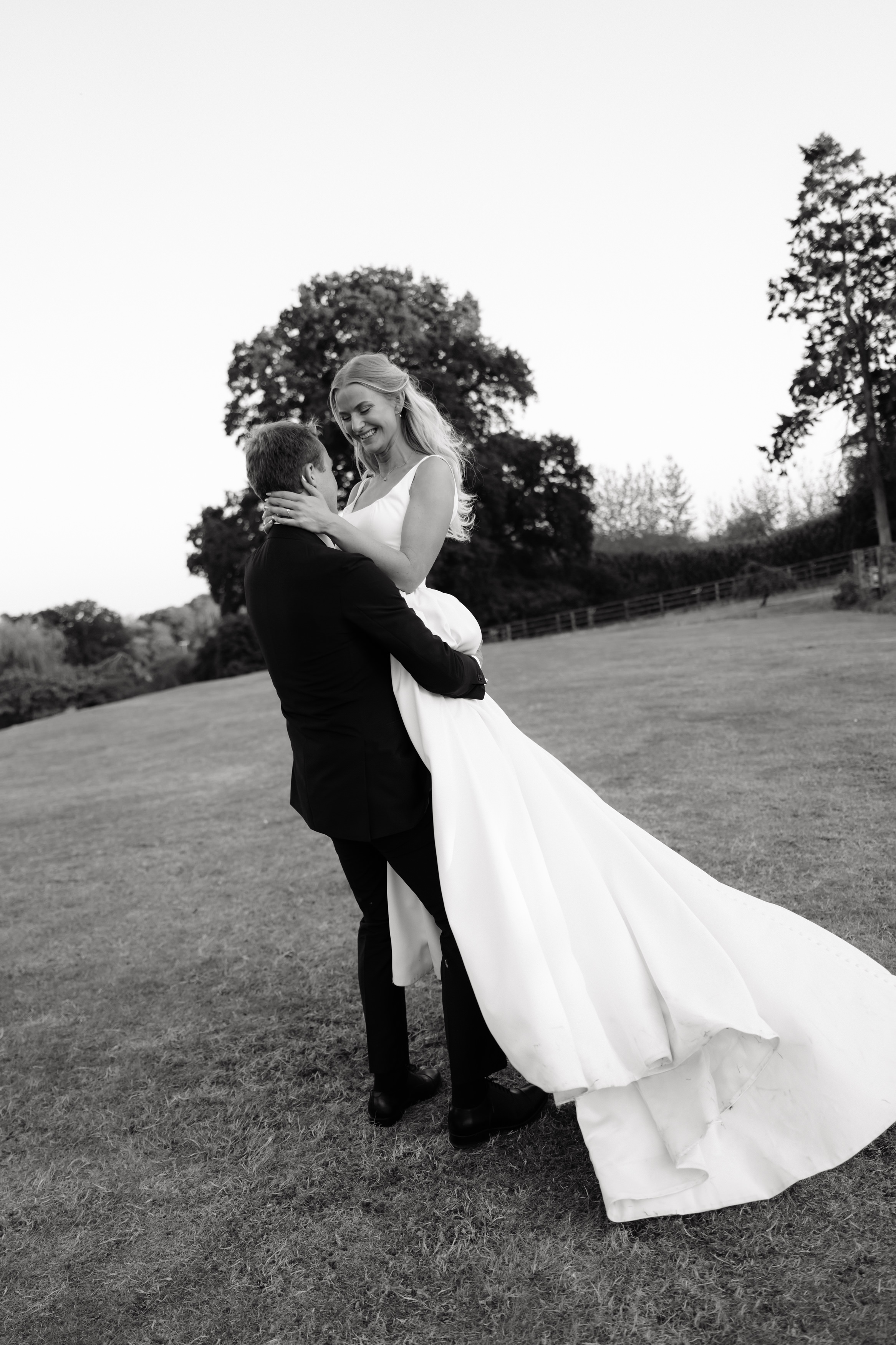 A man wearing a suit is picking up a woman in a wedding dress outside on the grass. They’re both smiling, and you can see trees in the background.