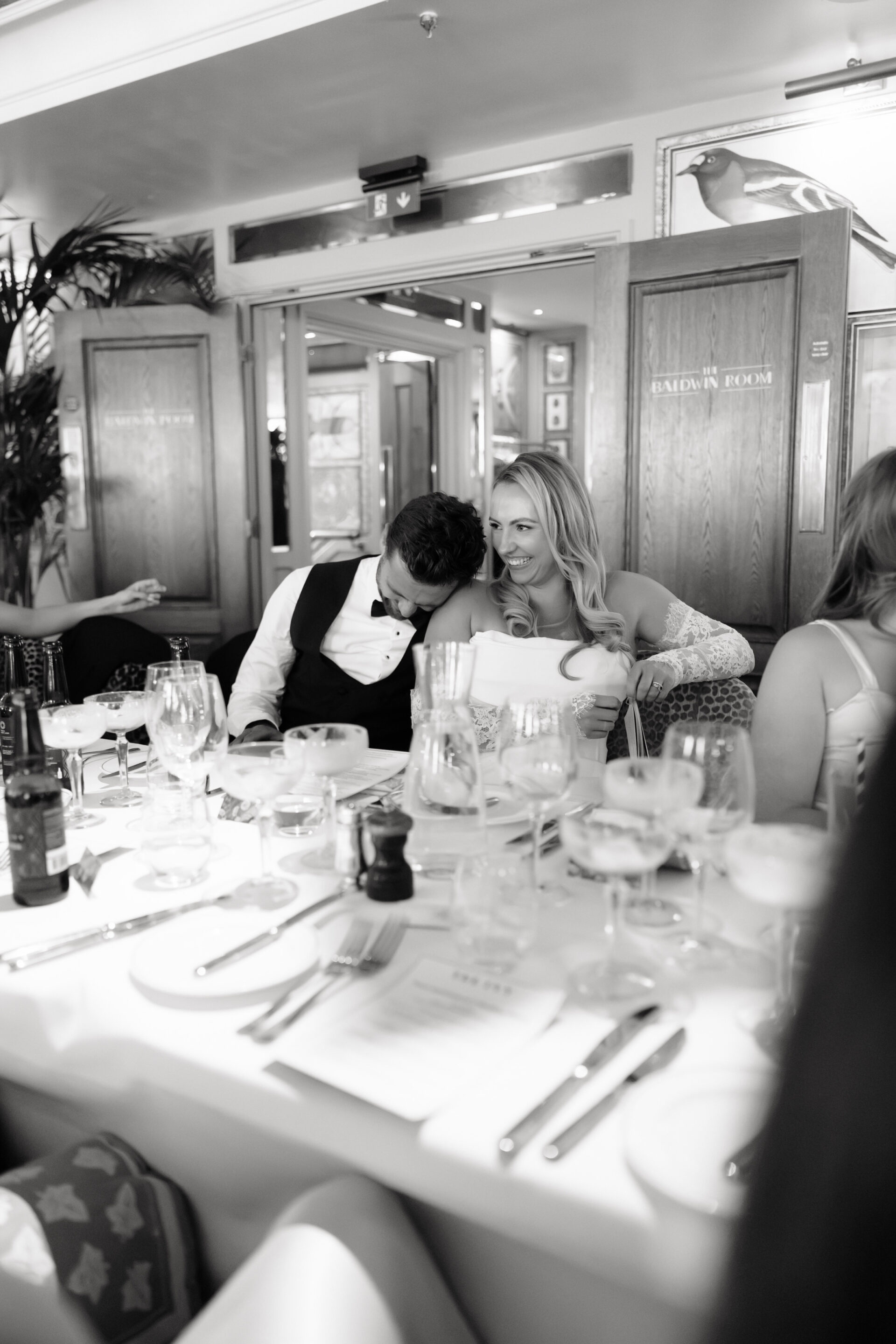 A man leans his head on a woman’s shoulder while they sit together at a smart dining table in a bright restaurant, with plates and glasses all around them.