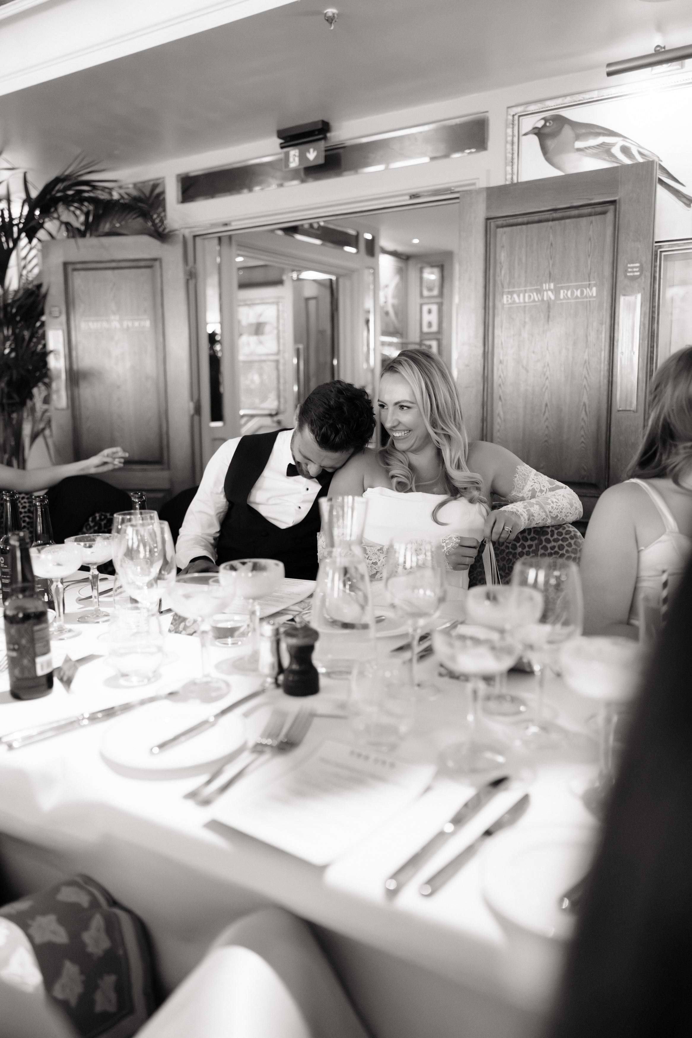 A man leans his head on a woman’s shoulder while they sit together at a smart dining table in a bright restaurant, with plates and glasses all around them.