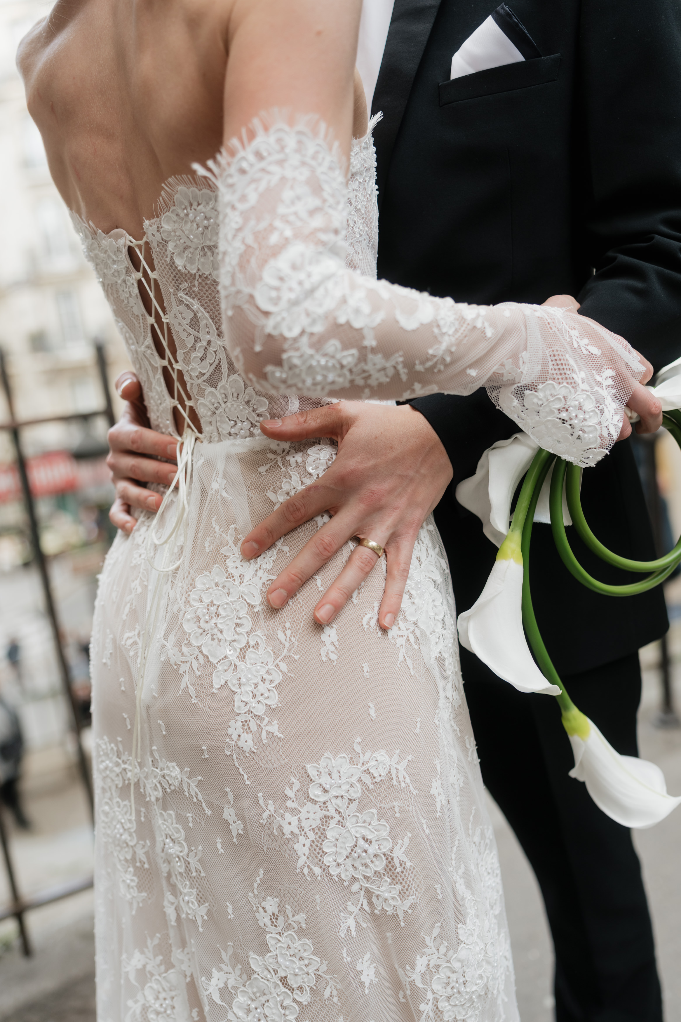 A bride in a lace dress stands next to a groom in a black suit. They have their arms around each other, holding white calla lilies.