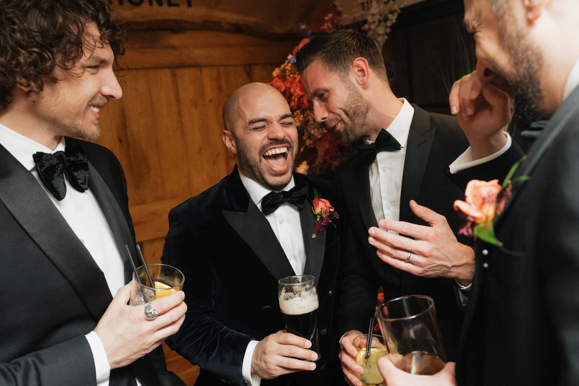 Four guys in tuxes are indoors, chatting and laughing with drinks in hand at a fancy event or party.
