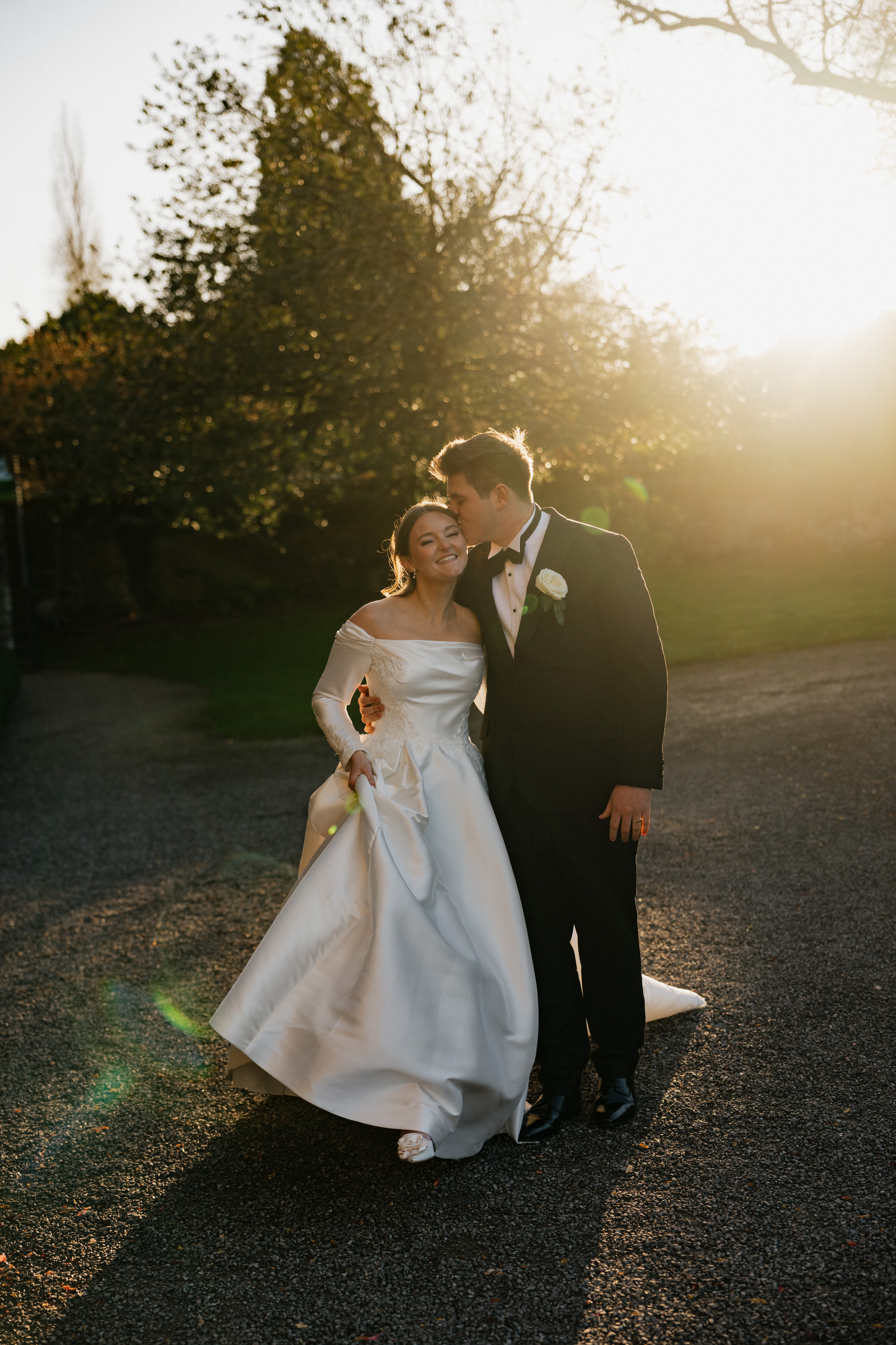 A bride in a white dress and her groom in a black tuxedo walk together at sunset, smiling and linking arms on a gravel path.