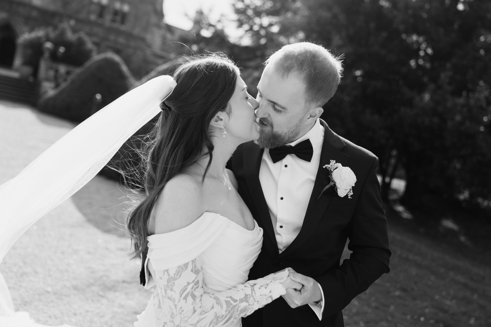 A bride and groom kiss outside, holding hands. She wears an off-shoulder dress and veil; he’s in a dinner suit with a flower pin.