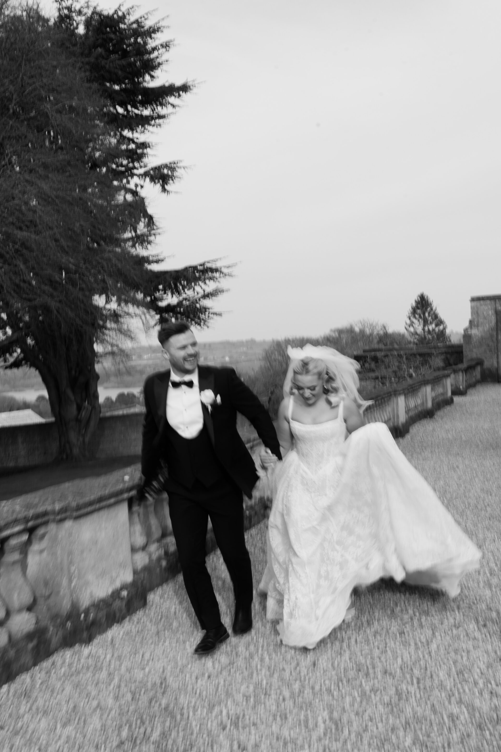 A bride and groom in wedding clothes walk briskly outside on a stone path, holding hands, with trees and a stone wall behind them.