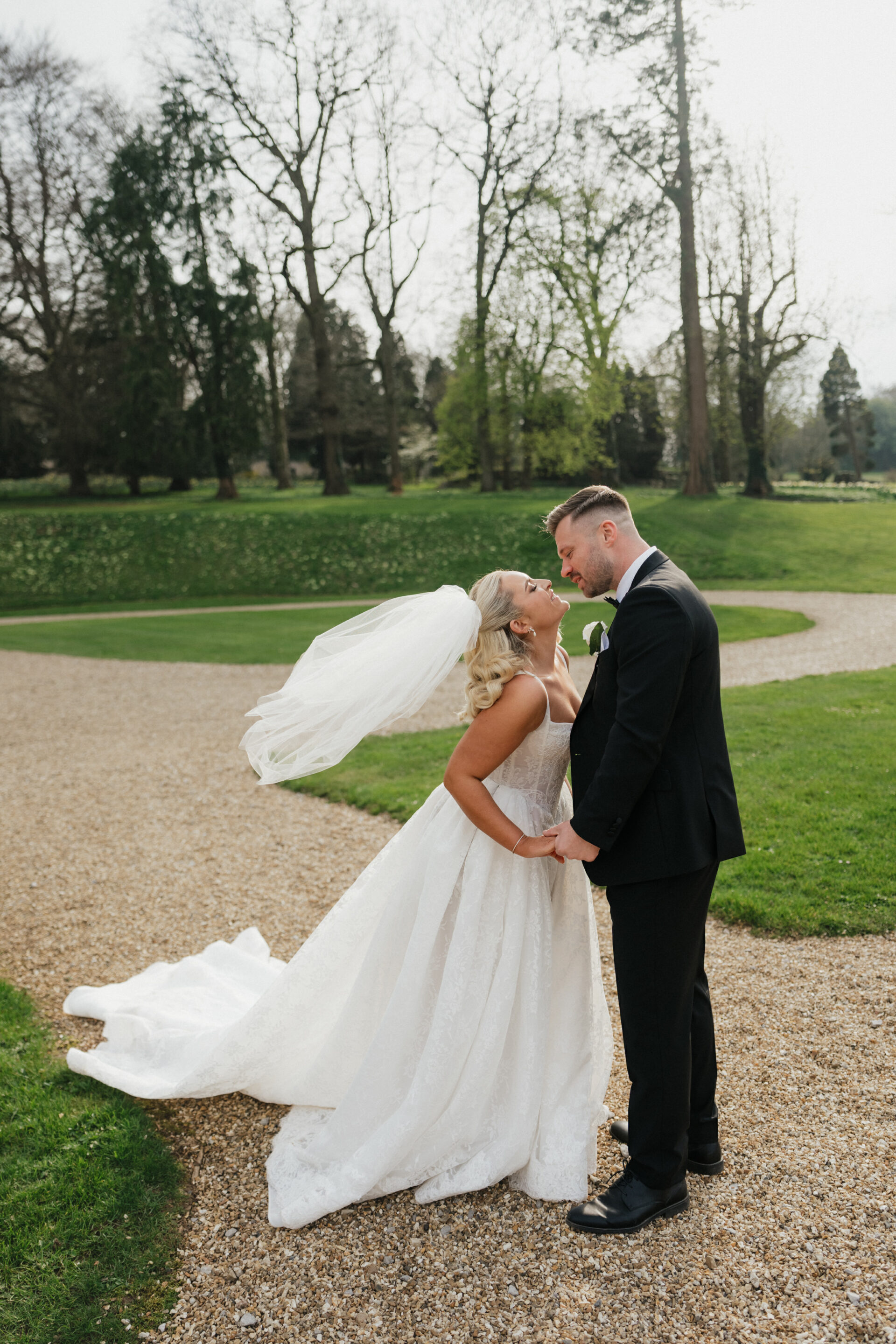 A bride and groom are outside on a gravel path, holding hands and smiling at each other, with grass and tall trees around them.