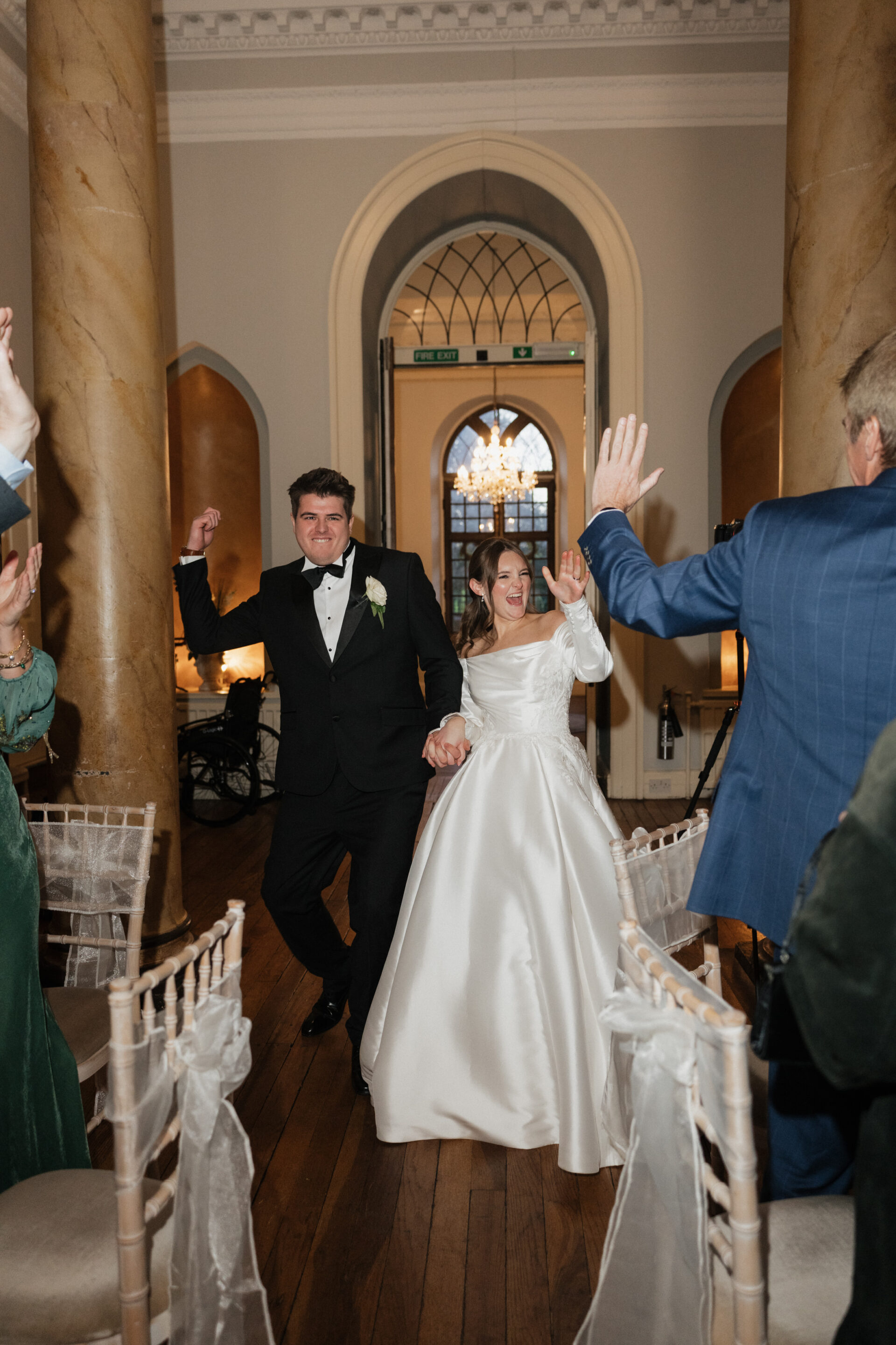 The bride and groom walk down the aisle holding hands, grinning and waving as their guests applaud in a posh hall with columns.