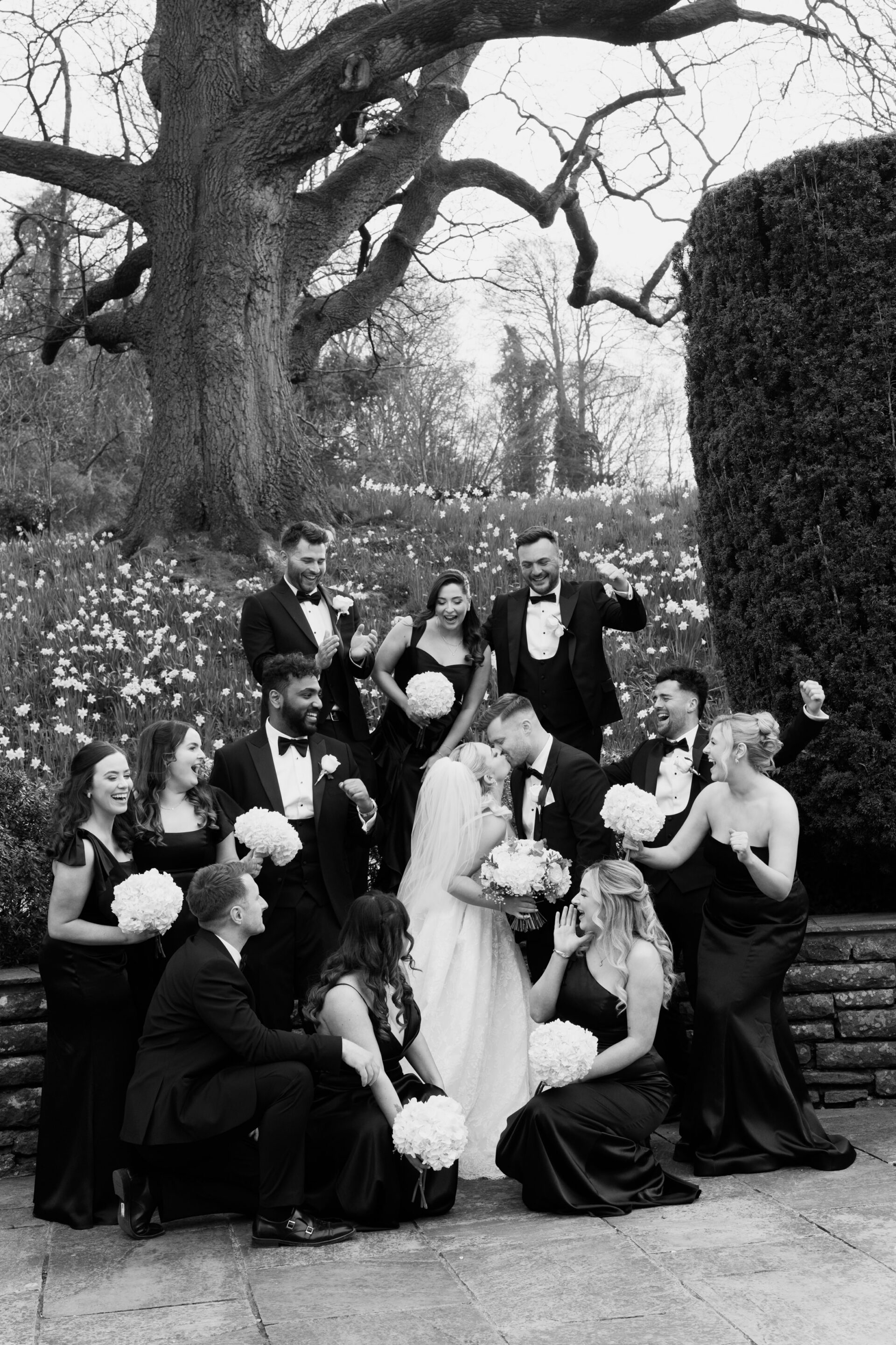 A bride and groom share a kiss outside, with their wedding party in black outfits next to a big tree and some shrubs.