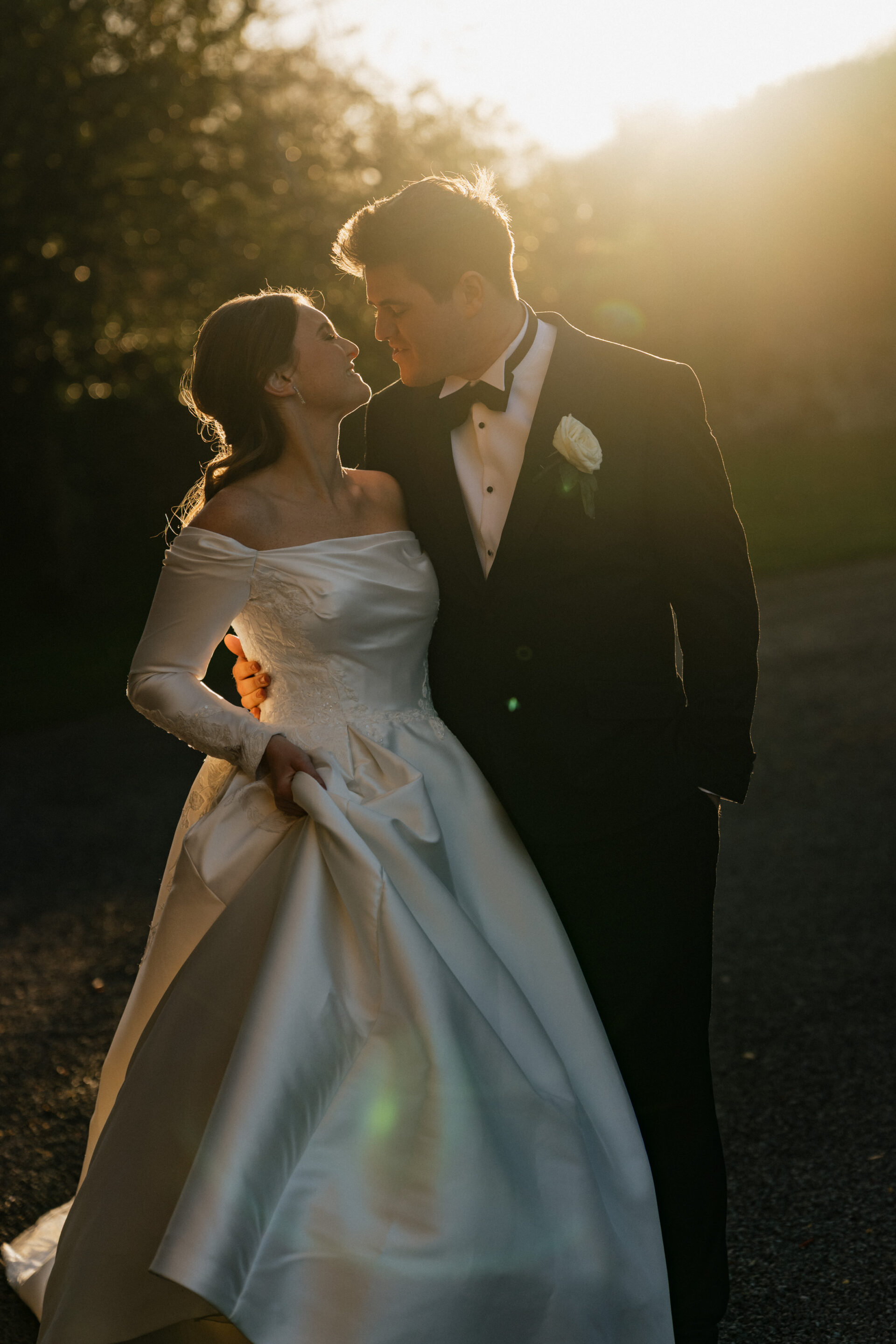 A bride in a white dress and a groom in a suit stand close together outside at sunset, gazing into each other’s eyes.
