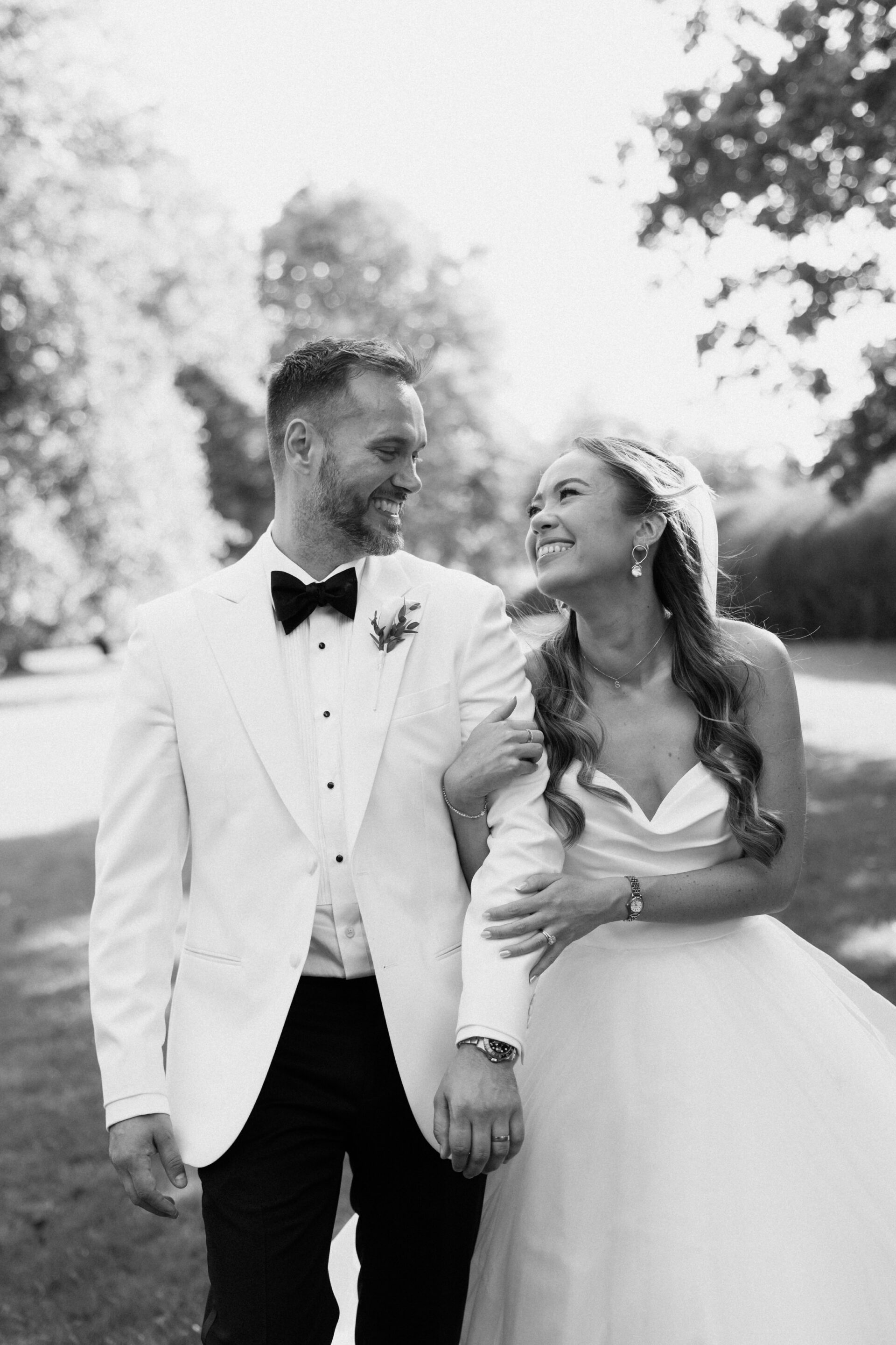A bride and groom walk outside together, dressed up for their wedding, smiling as they look at each other.
