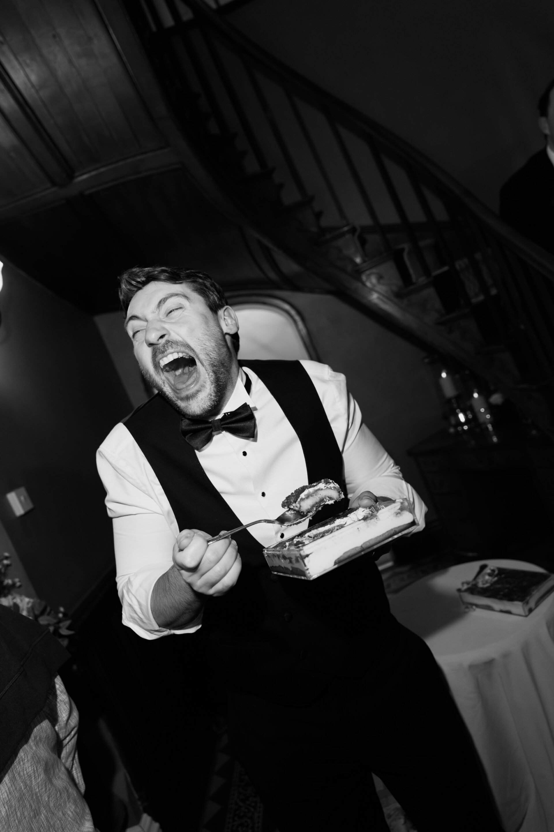 A man dressed up is laughing and holding a plate of cake and a fork at an indoor event, with stairs behind him.