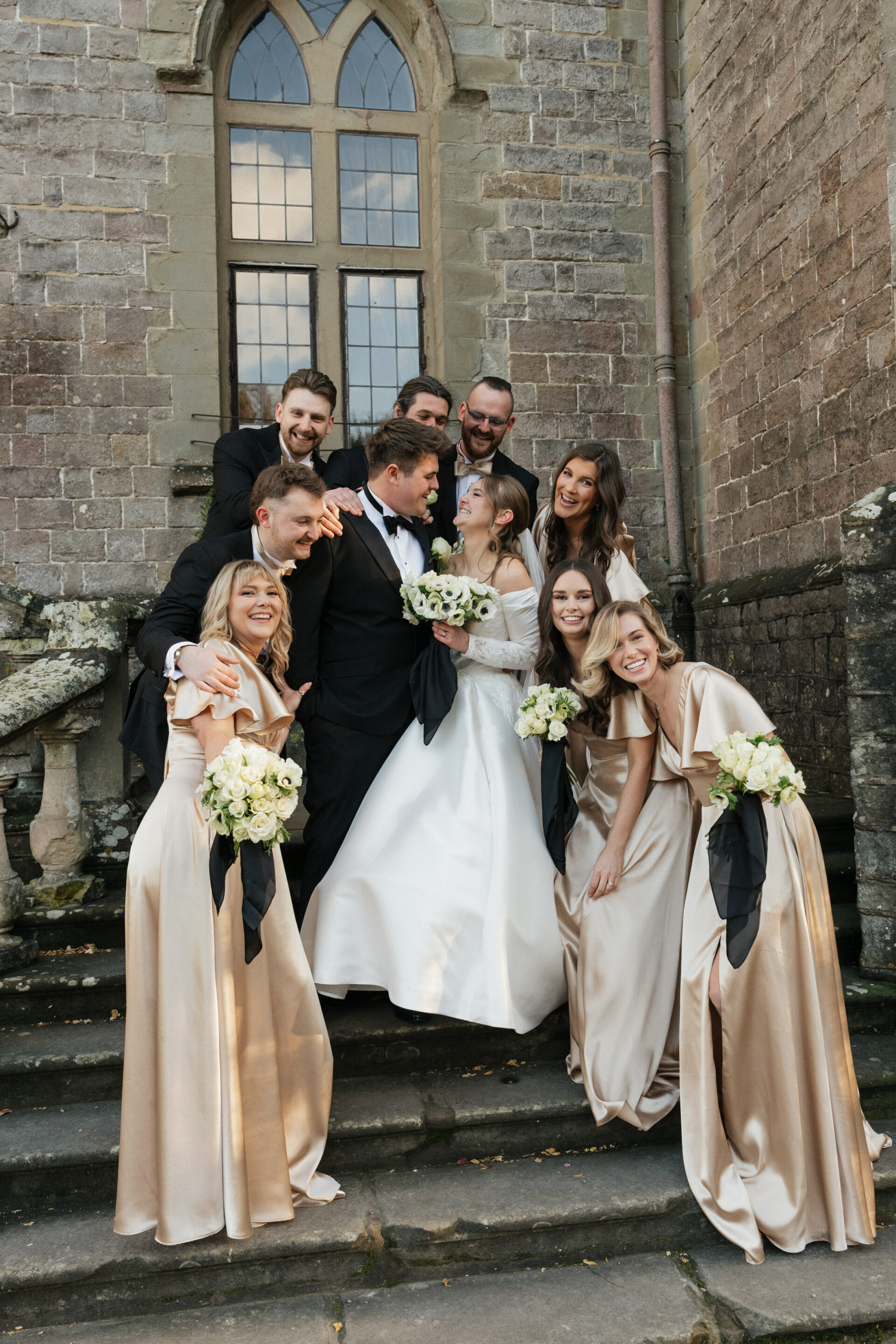 The bride and groom are on stone steps with their friends in dresses and suits, all happily smiling outside a stone building.