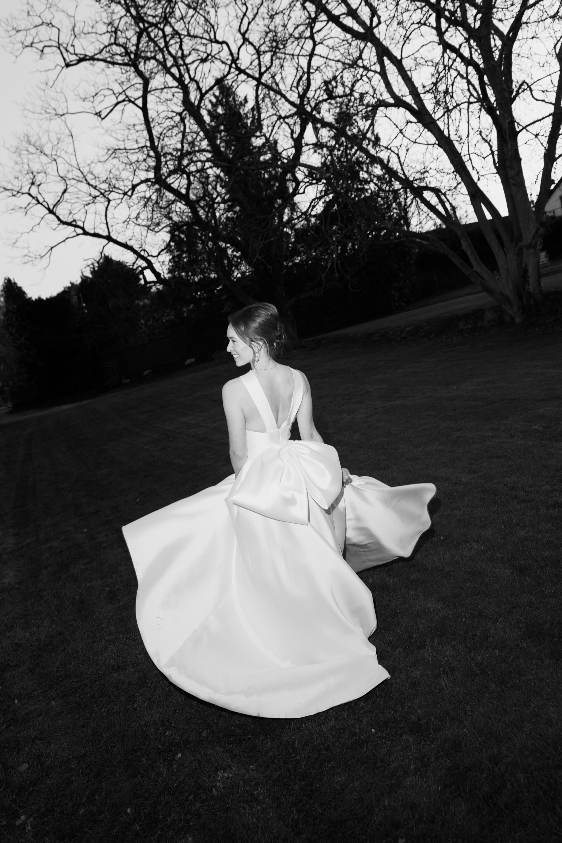 A woman wearing a white dress with a large bow on the back stands on grass, facing away, with bare trees and bushes behind her.
