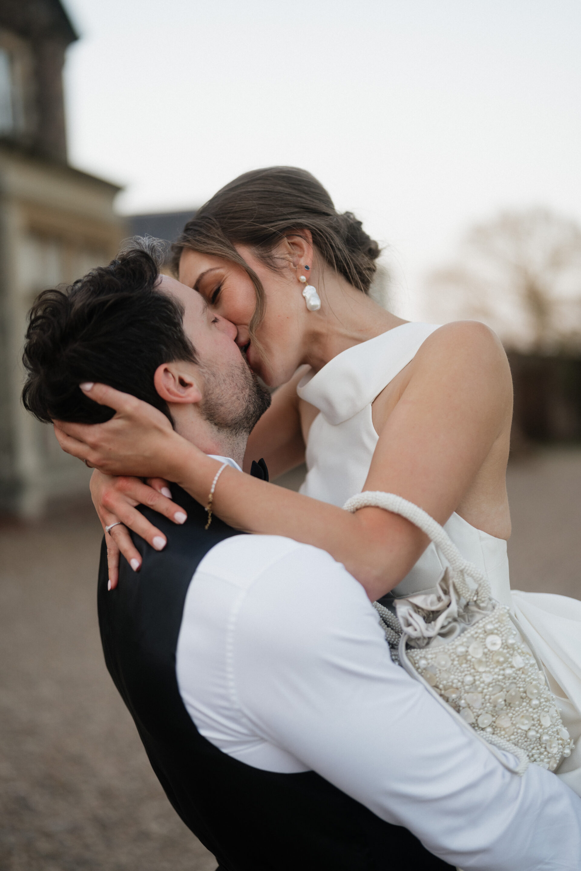 A bride and groom share a kiss outside. He’s holding her up, she’s in a white dress, and he’s wearing a white shirt with a dark waistcoat.