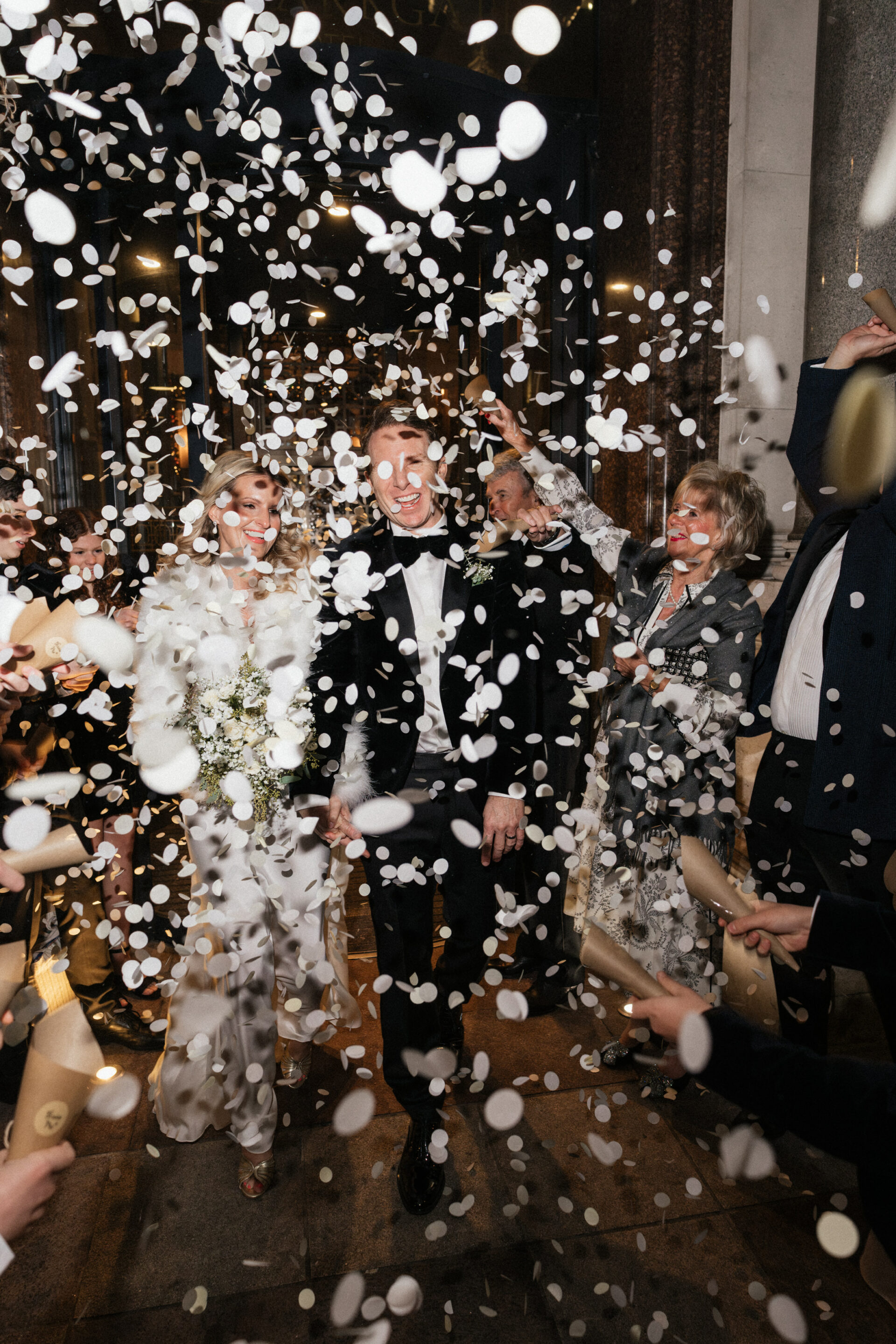 A bride and groom, dressed up for their wedding, walk through a crowd at night as guests throw white confetti around them.