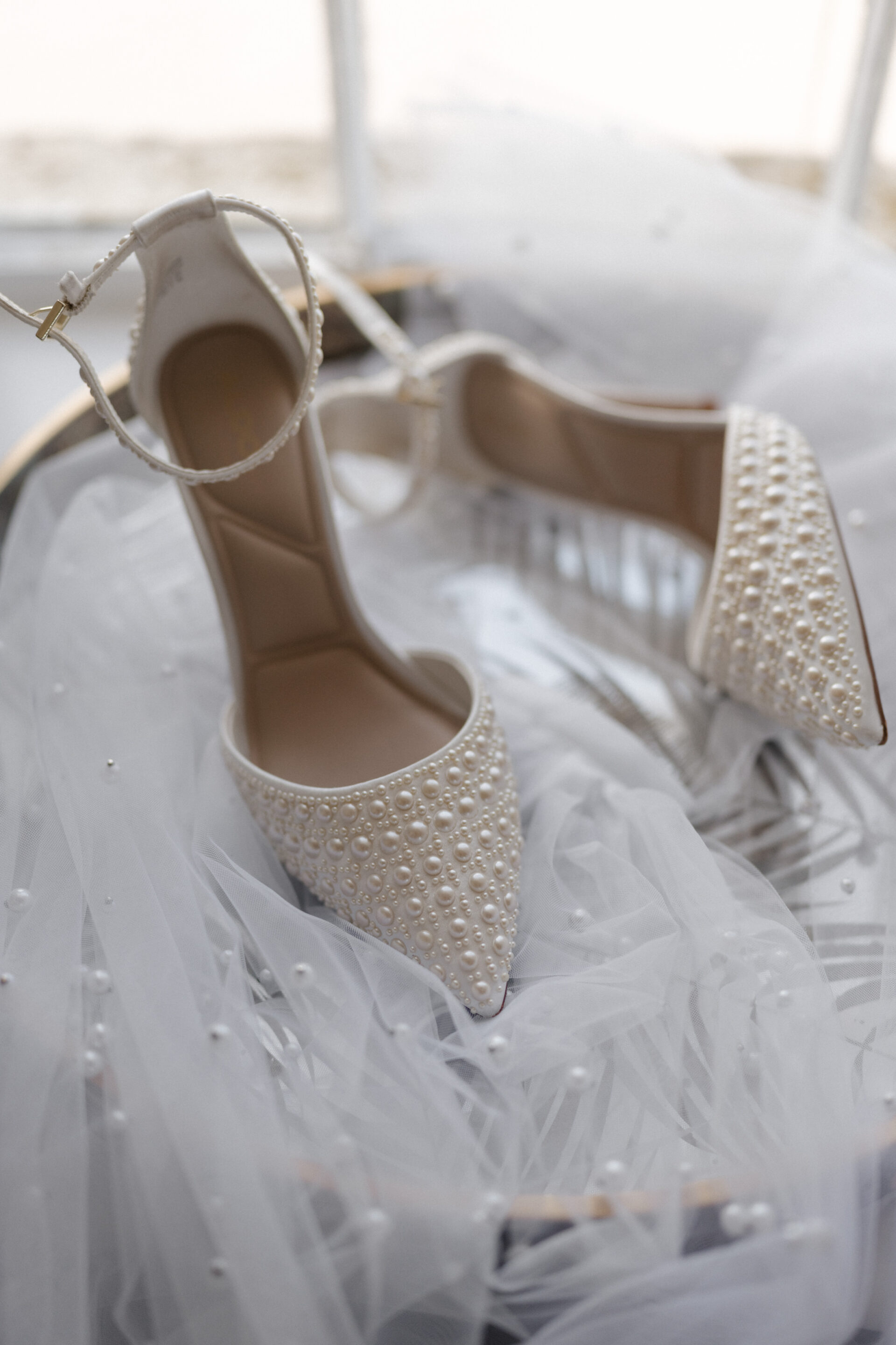 White high heels with ankle straps and pearls sit on see-through fabric with pearls, near a window.
