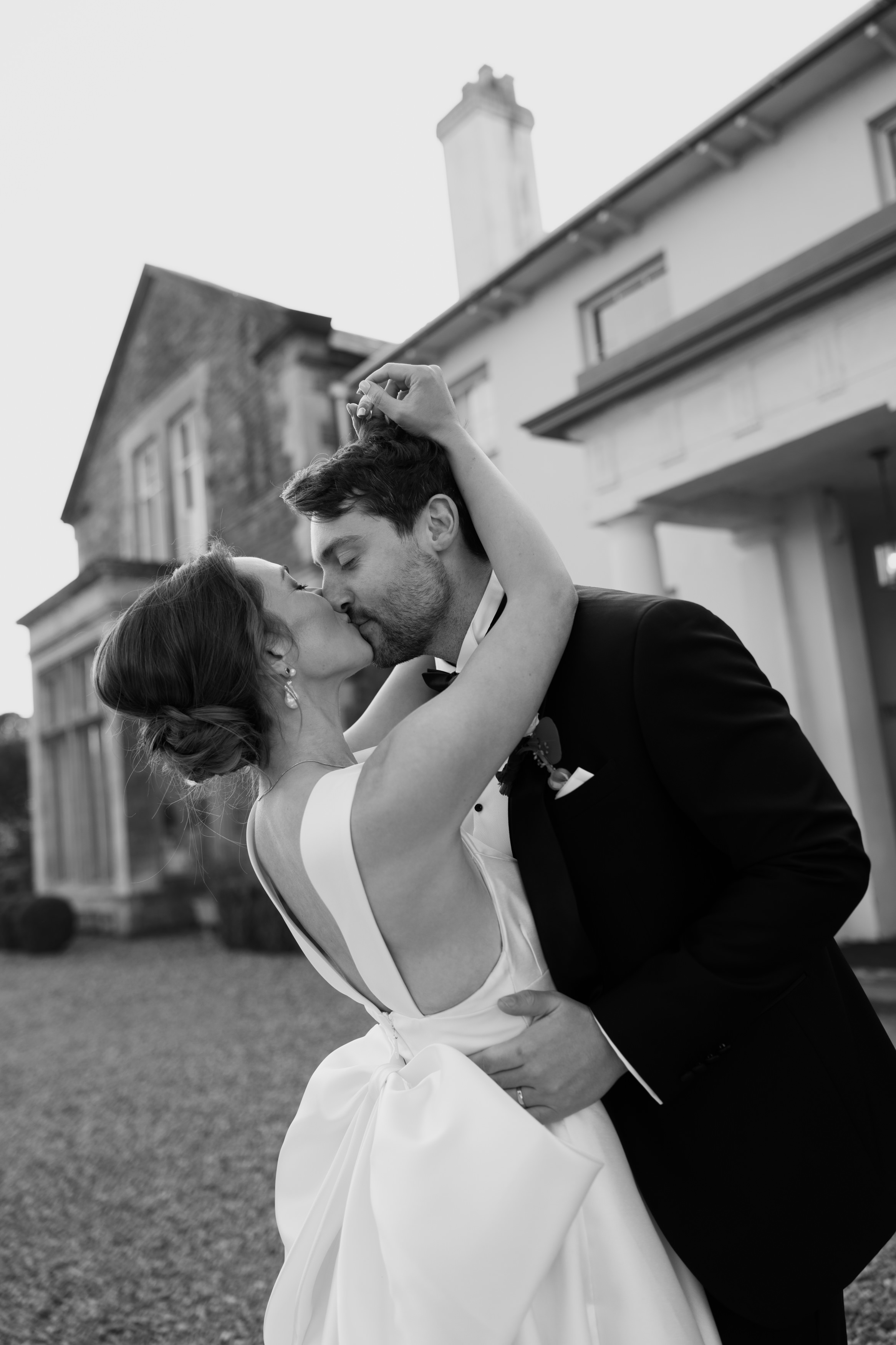 A bride and groom kiss outside near a large building, the bride's back faces the camera as she lifts her hand.