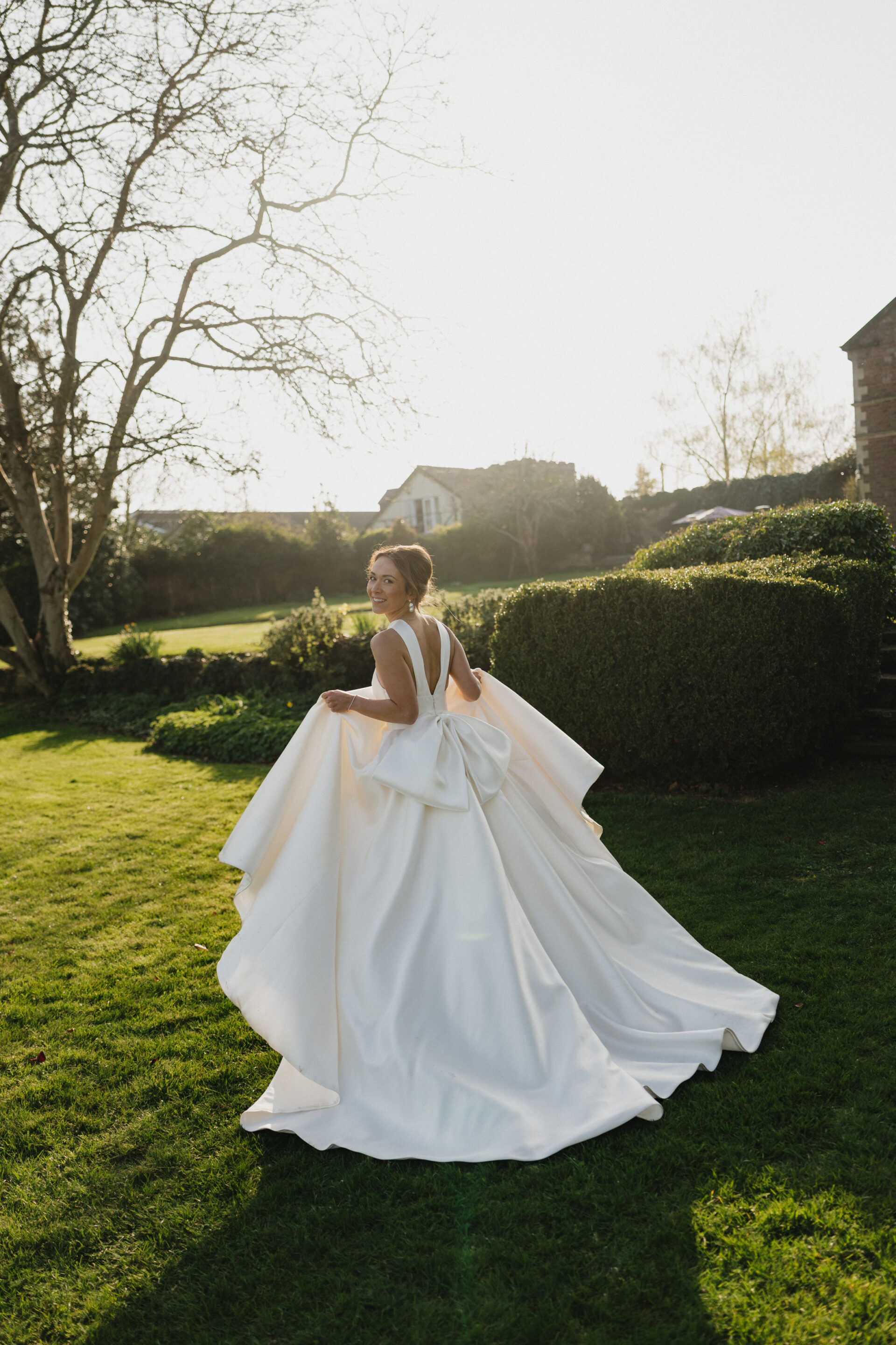 A woman in a white wedding dress stands outside on the grass, holding her dress and looking back. Trees and a house are behind her.