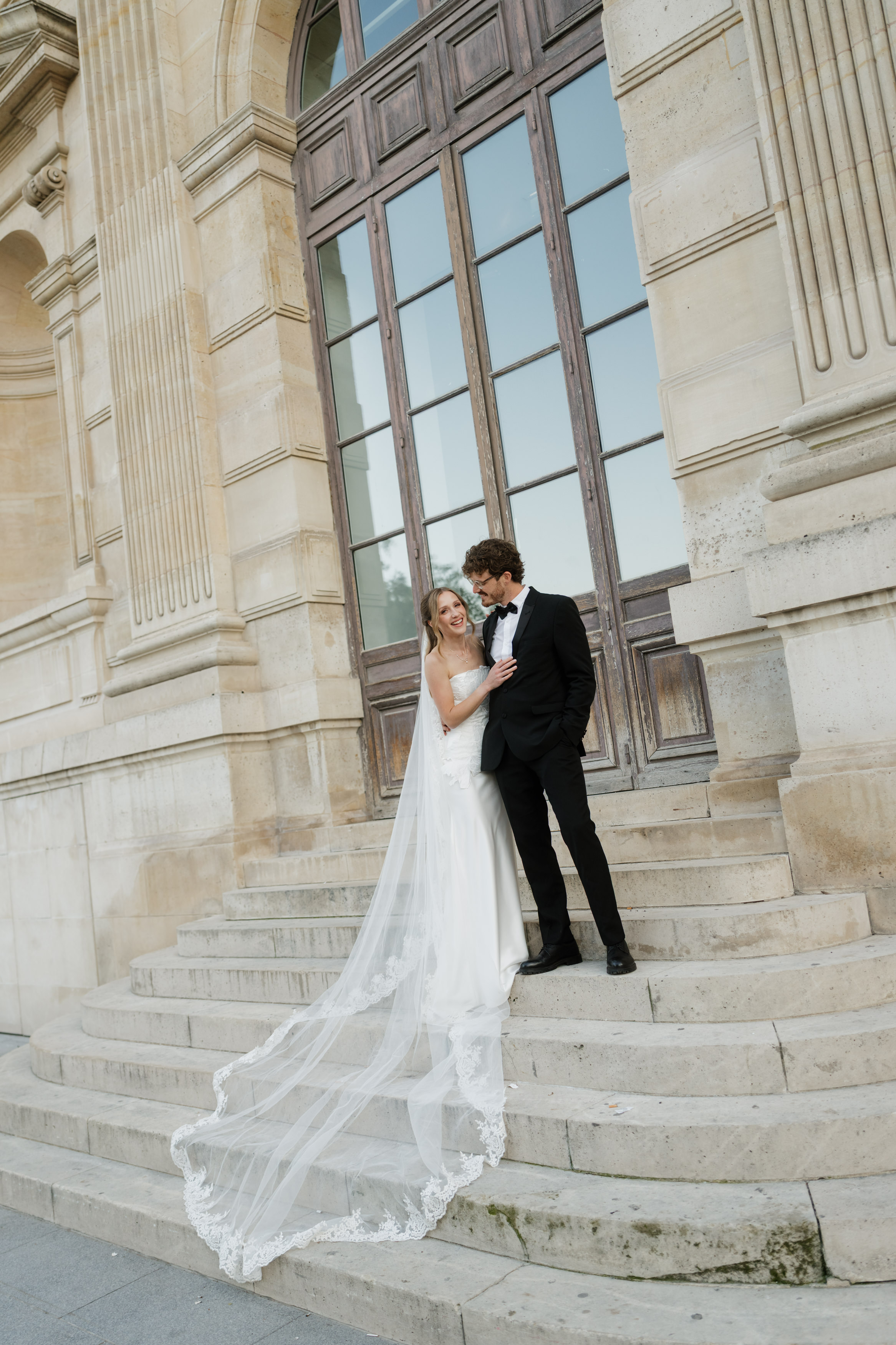 A bride and groom are on stone steps by a big arched wooden door, and the bride’s long veil trails down the steps.